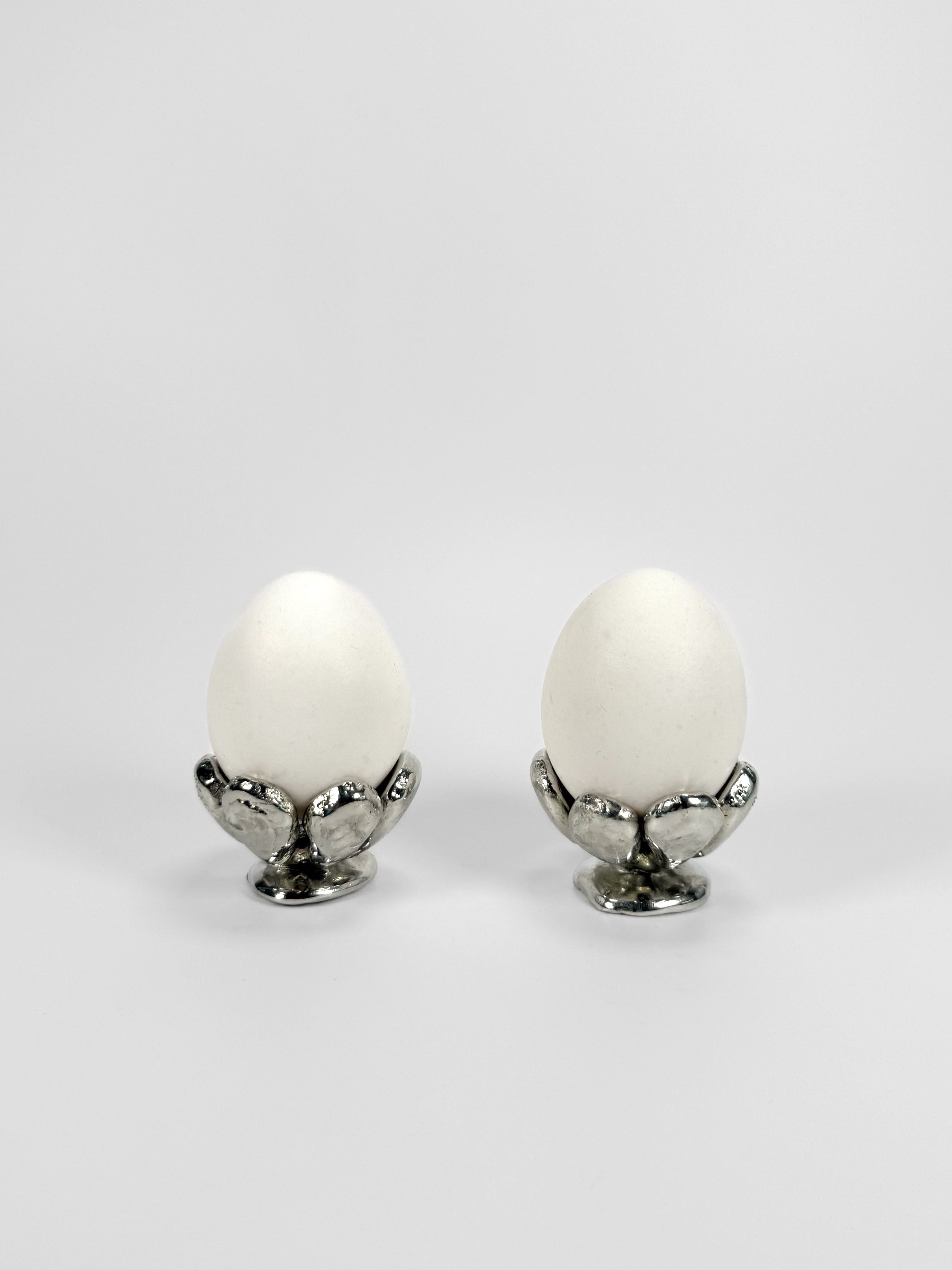 Two white eggs sit in Studio Formbart's The Tin Collection / FLWR egg cups, shaped like flower petals, against a plain white background.