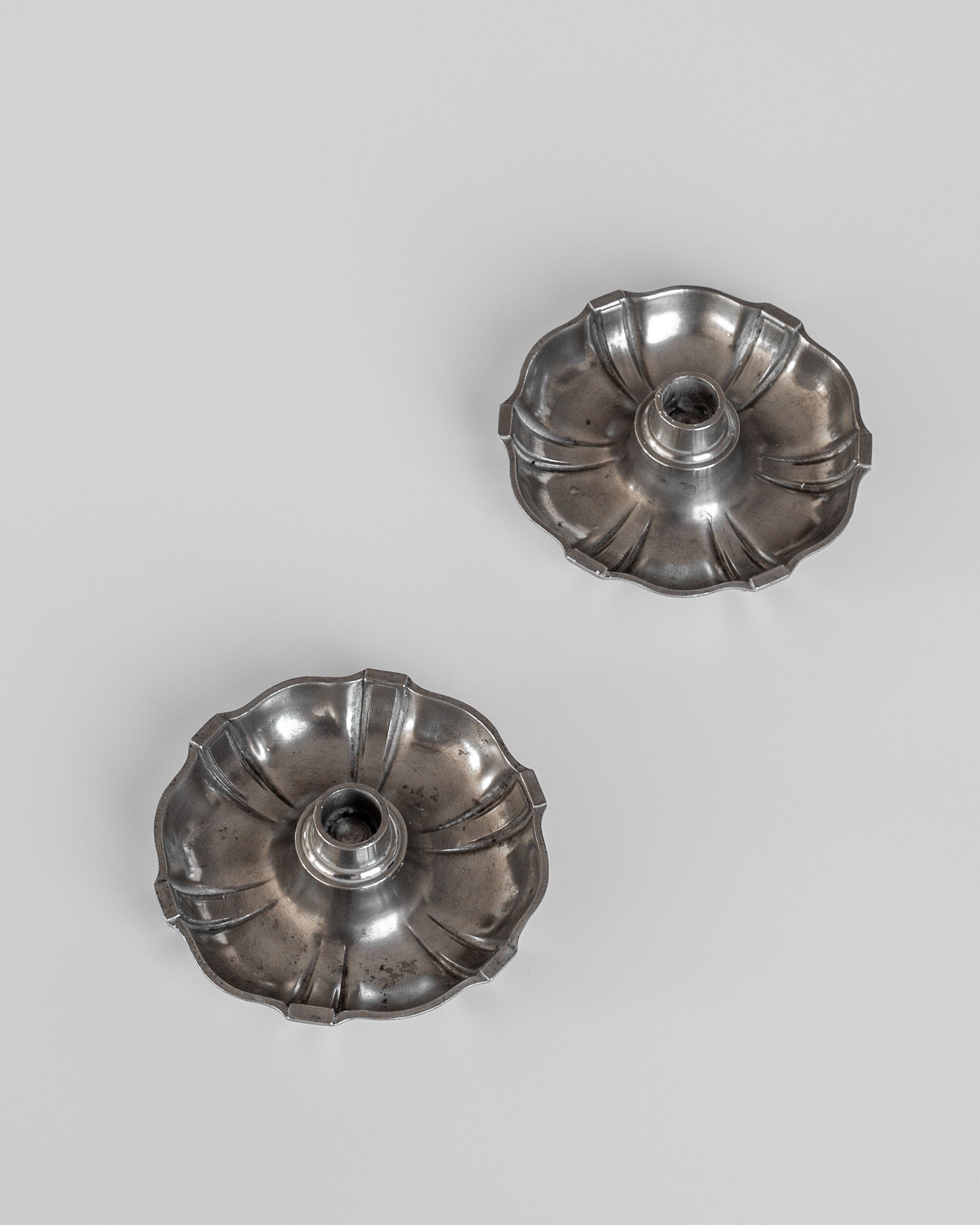 Two sophisticated Spigel Modernist Metal Candleholders from the 1930s, with scalloped edges, grace a plain gray background. Emblematic of Swedish Modernist metalwork and reminiscent of C.G. Hallberg's craftsmanship, these pieces include a central socket for candle placement.