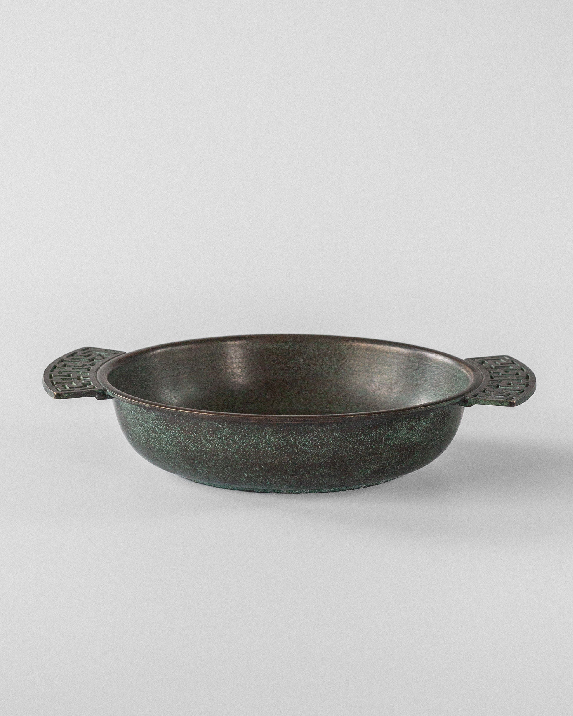 The Spigel Art Deco Bronze Bowl, from 1930s Sweden, is a round, texturally charming pot with dark green metal and ornate Greek key handles on each side, placed on a plain white background.