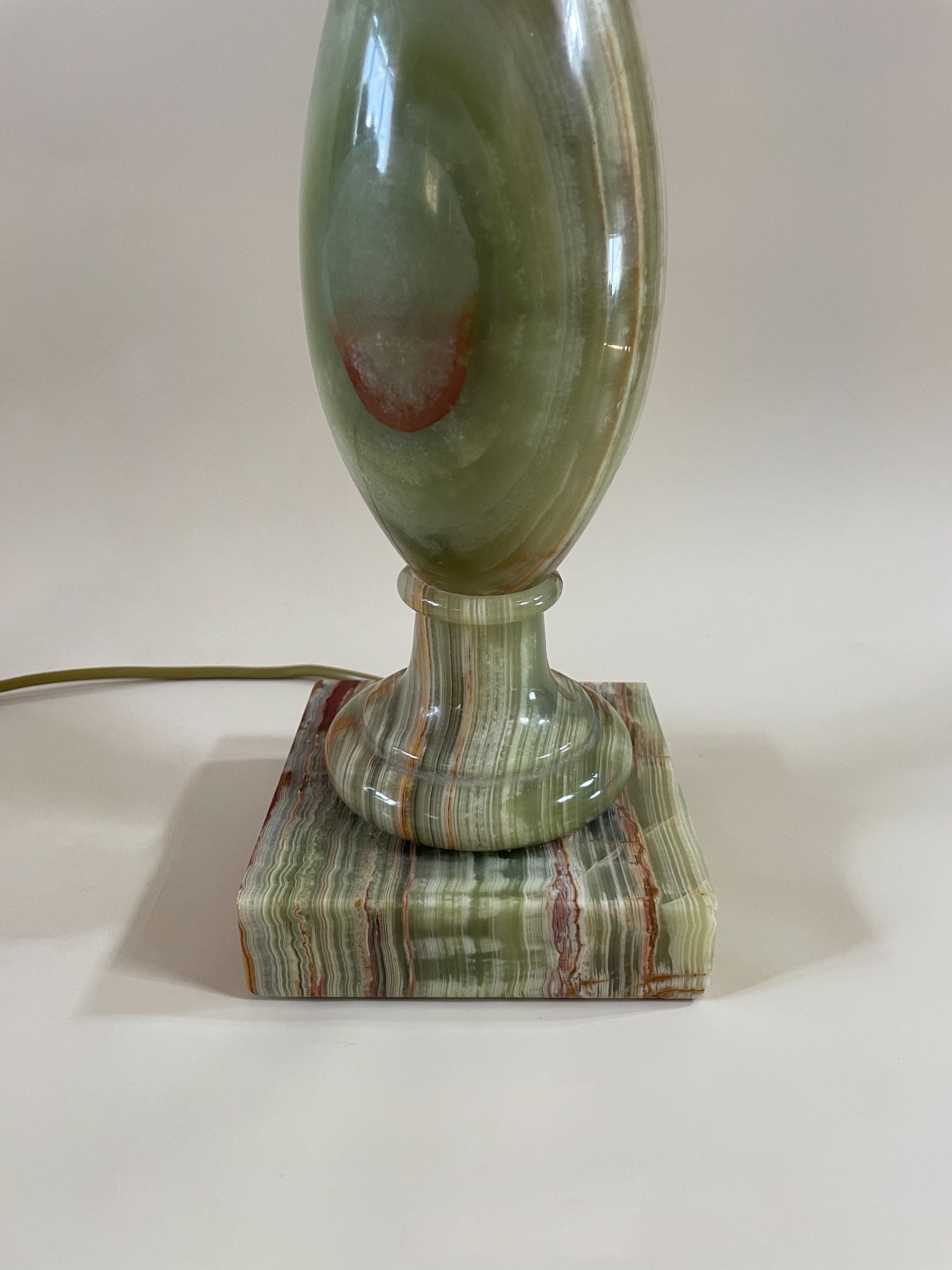 A close-up image of the Green Onyx Lamp C.1970 by Maud Vaughan, showcasing its vintage charm with a smooth, polished surface. The lamp features a rounded base and an elongated, oval-shaped body, all resting on a square, striped onyx platform. A power cord is visible extending from the back of the lamp.