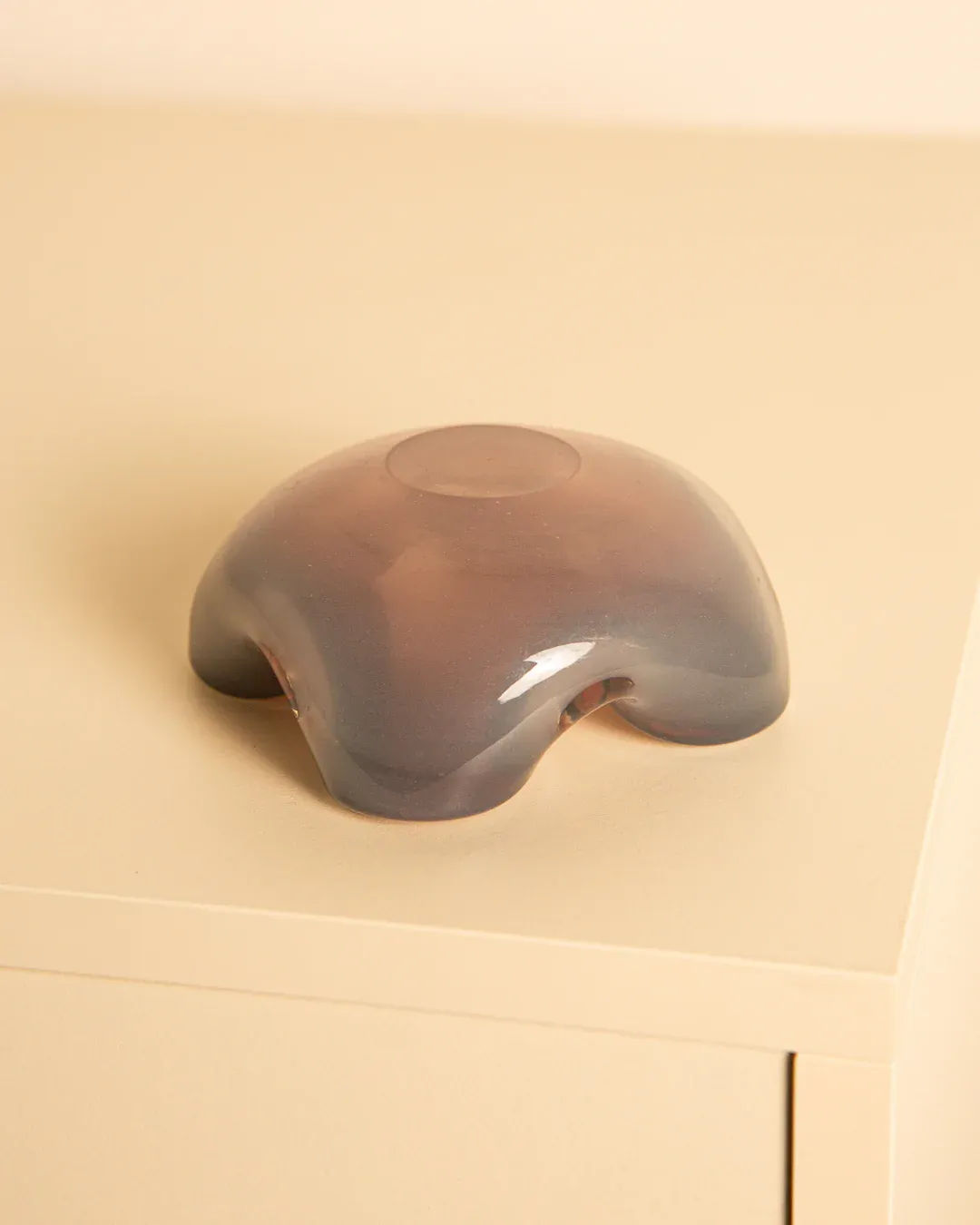 The Treaptyque Murano Opaque Glass “Fleur” 60s, with its smooth, dome-shaped design and four rounded protrusions, sits elegantly on a light wooden surface. Its muted pink to gray gradient evokes the artistry of vintage Italian glassmakers.