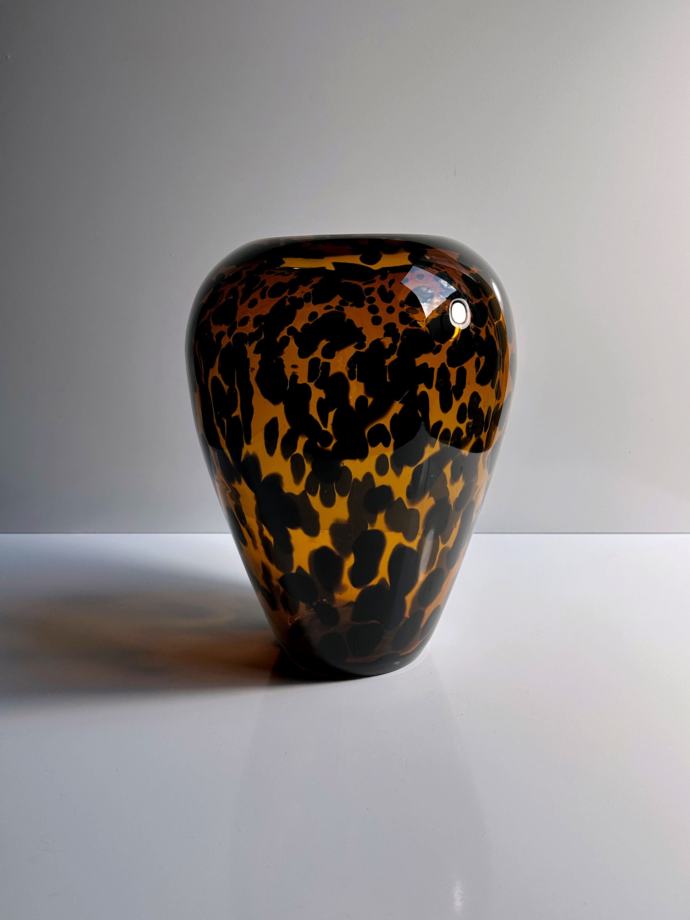 The Tortoise vase Murano from the 1960s, offered by Dodo Vintage for Christian Dior, showcases a striking tortoiseshell pattern of dark brown and tan spots against a white background. Its rounded, glossy surface elegantly reflects light, enhancing its polished and timeless chic appearance.