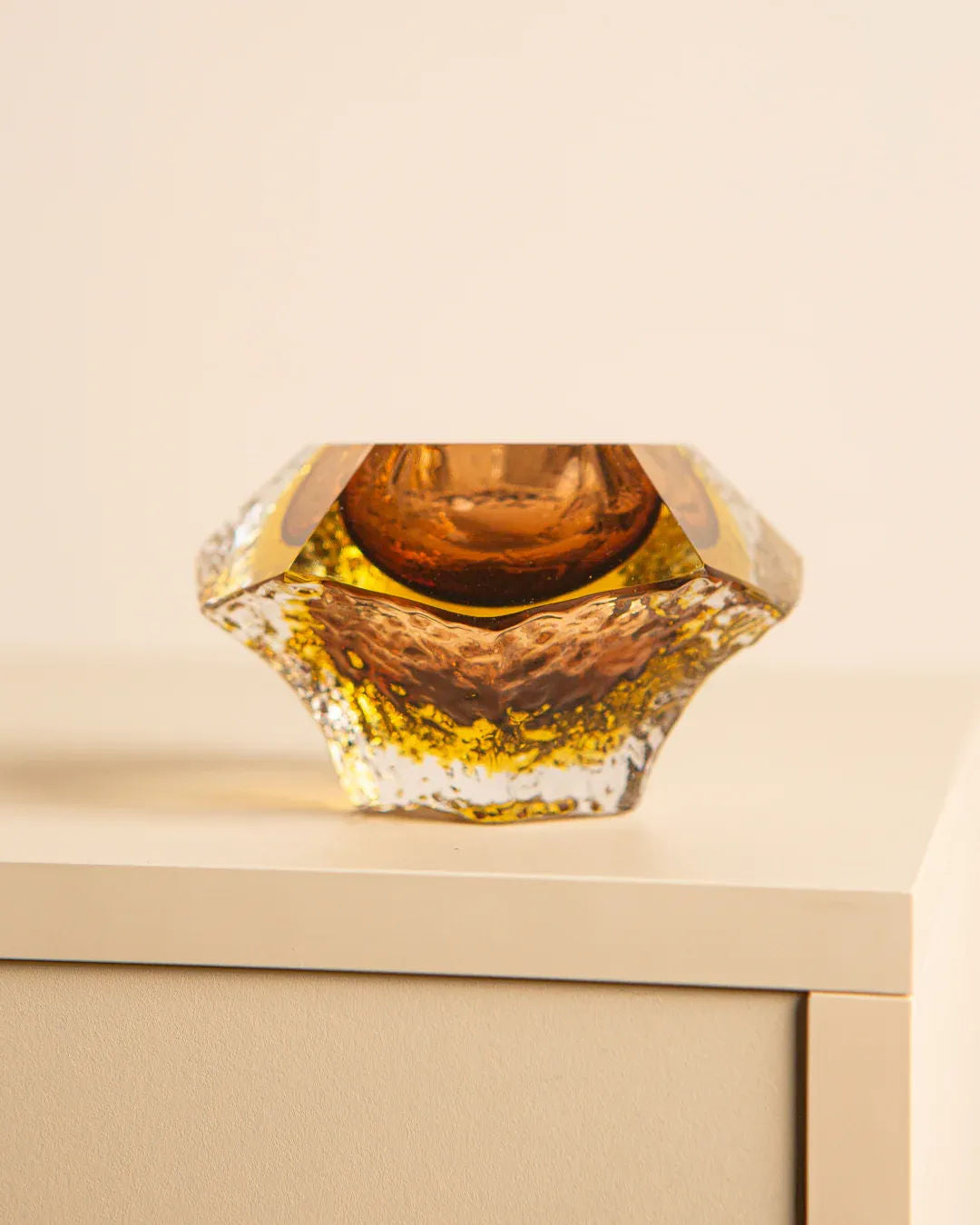 Resting on a light-colored, minimalist surface, the 'Diamant' ashtray by Flavio Poli for Seguso 60’s from Treaptyque echoes Italian design with its geometric amber glass, textured star-like shape, subtle color variations, and elegant golden highlights.