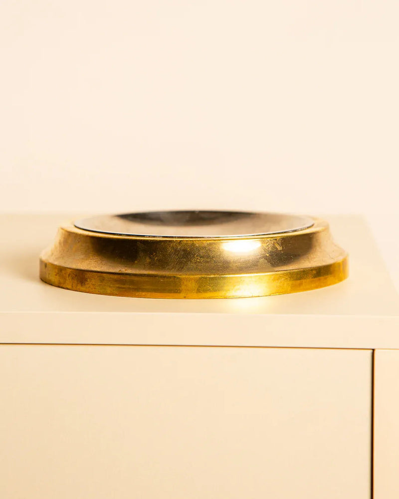 The Vide-poches in Bras Metal 60's by Treaptyque is a metallic, gold-toned circular piece with a reflective center that exudes vintage charm. Its sophisticated design rests elegantly on a light surface against a plain background, merging elegance with classic Italian allure.
