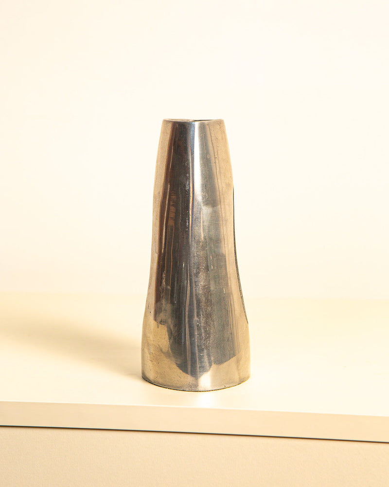 On a light-colored table against a cream background stands the tall, minimalist "Italian Vase in Aluminum 80's" by Treaptyque. Featuring a smooth, reflective surface and an elegantly simple shape with a slight taper, this design hails from Brescia, Italy.