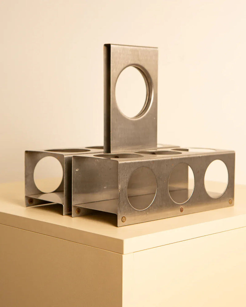 A Brushed Steel Wine Rack by François Monnet 70's, from the Treaptyque brand, with a rectangular base and four vertical panels featuring circular cutouts, is placed on a beige surface. The panels intersect at the center, creating a symmetrical structure with geometric shapes against the light brown background.