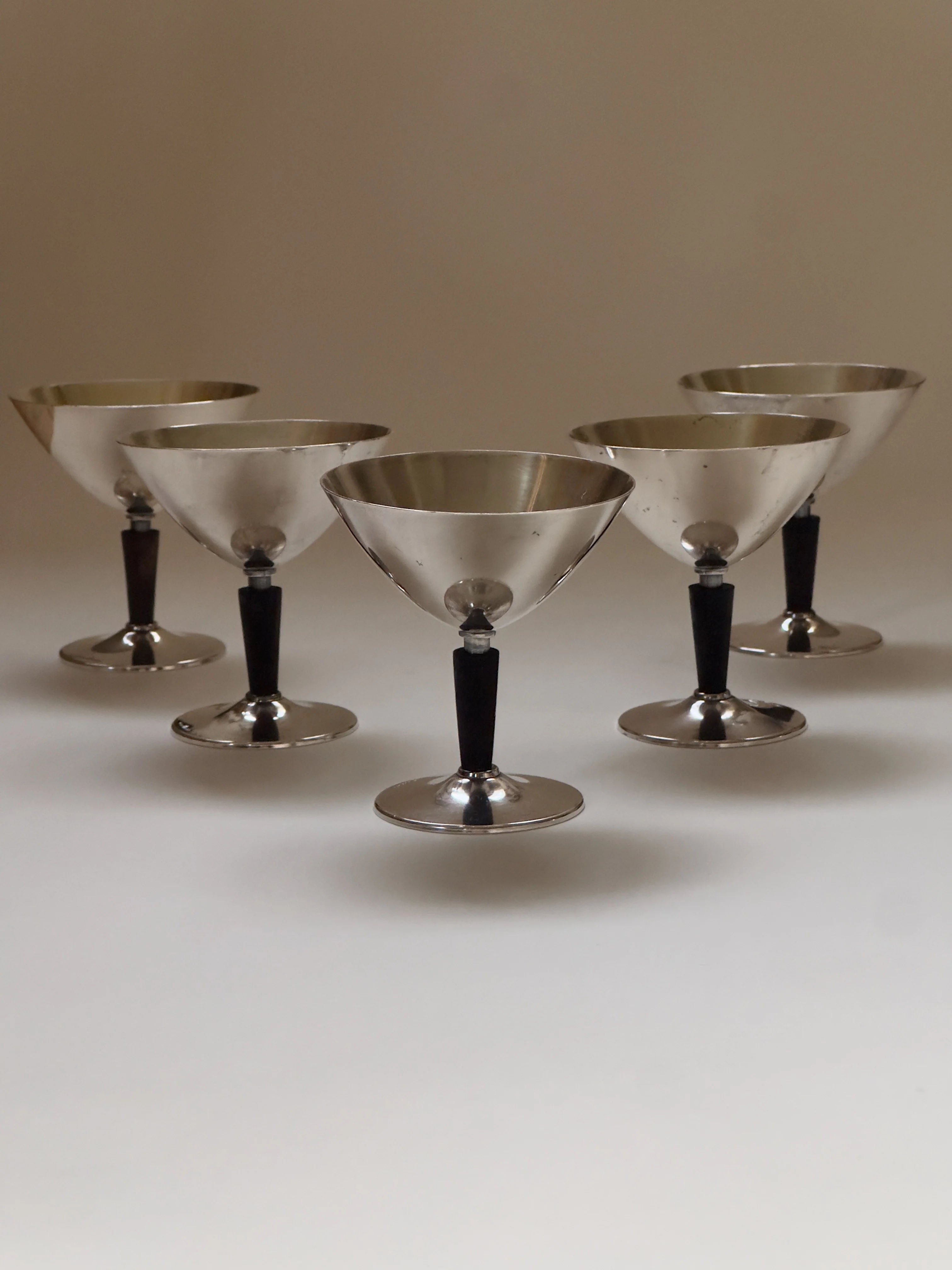 Five GAB Cocktail Glasses by Galerie Storm, made of vintage Swedish silver with black stems, are arranged in a staggered formation on a plain surface. The gleaming metal contrasts with the dark stems, exuding an elegant, minimalist look reminiscent of 1930s cocktail glasses.