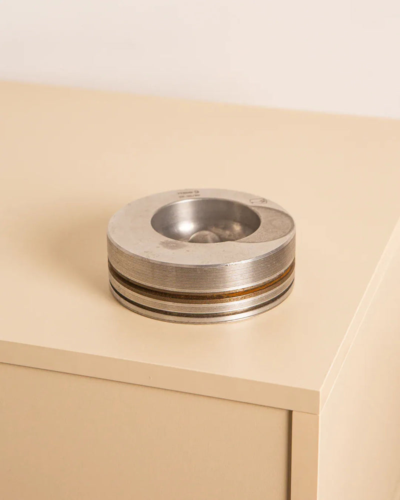 The Italian "Piston" ashtray in 80's metal by Treaptyque exudes vintage charm with its round, metallic design and features a transparent dome filled with small stones. It sits on a beige, minimalist desk against a light background, adding an elegant touch reminiscent of Milanese design.