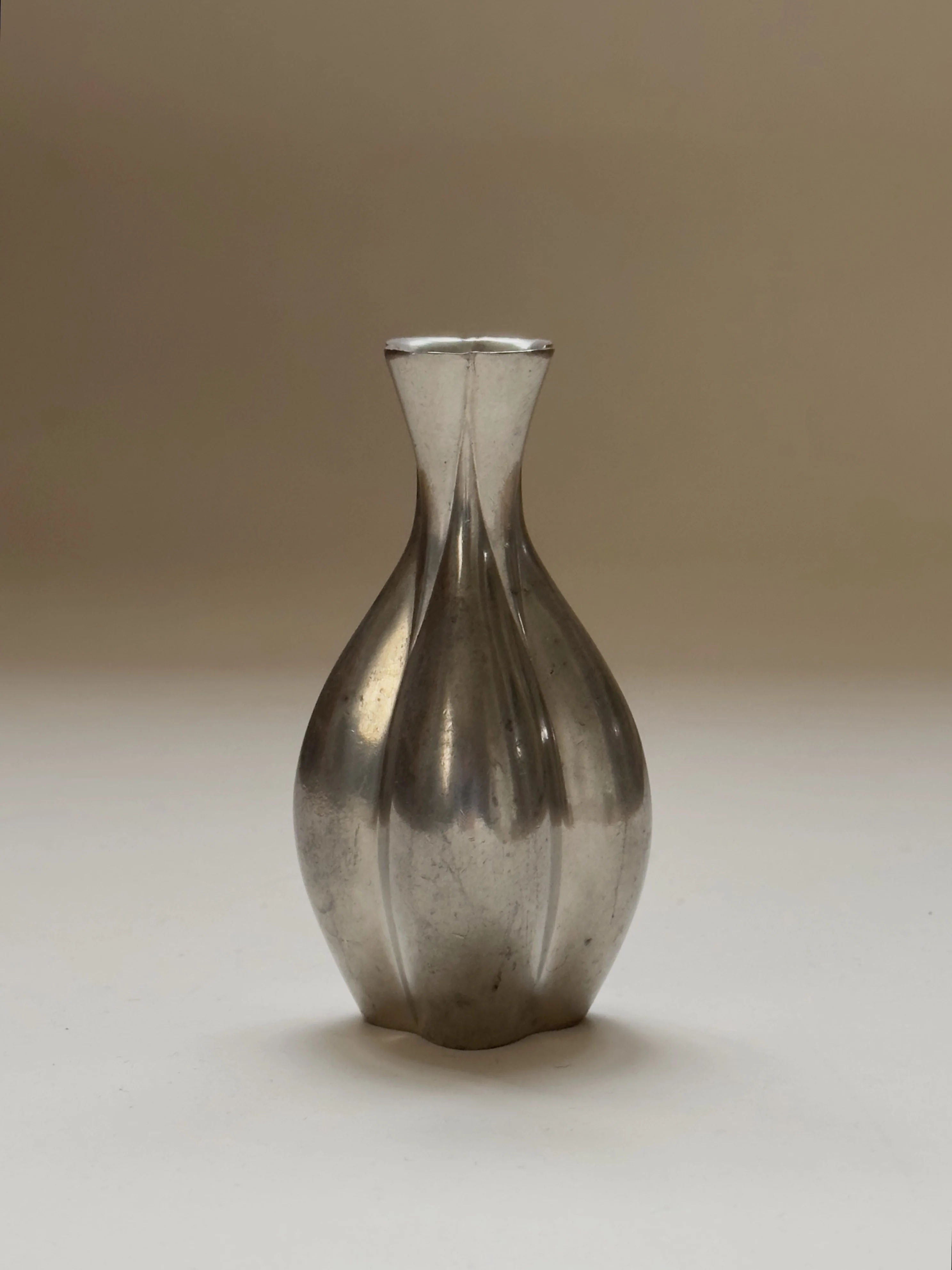 This Galerie Storm Just Andersen Pewter Vase, with its vintage 1940s charm, showcases a silver, vase-shaped design and smooth, reflective surface. Vertical ridges enhance its elegance. It has a bulbous base tapering to a small opening against a plain beige backdrop.