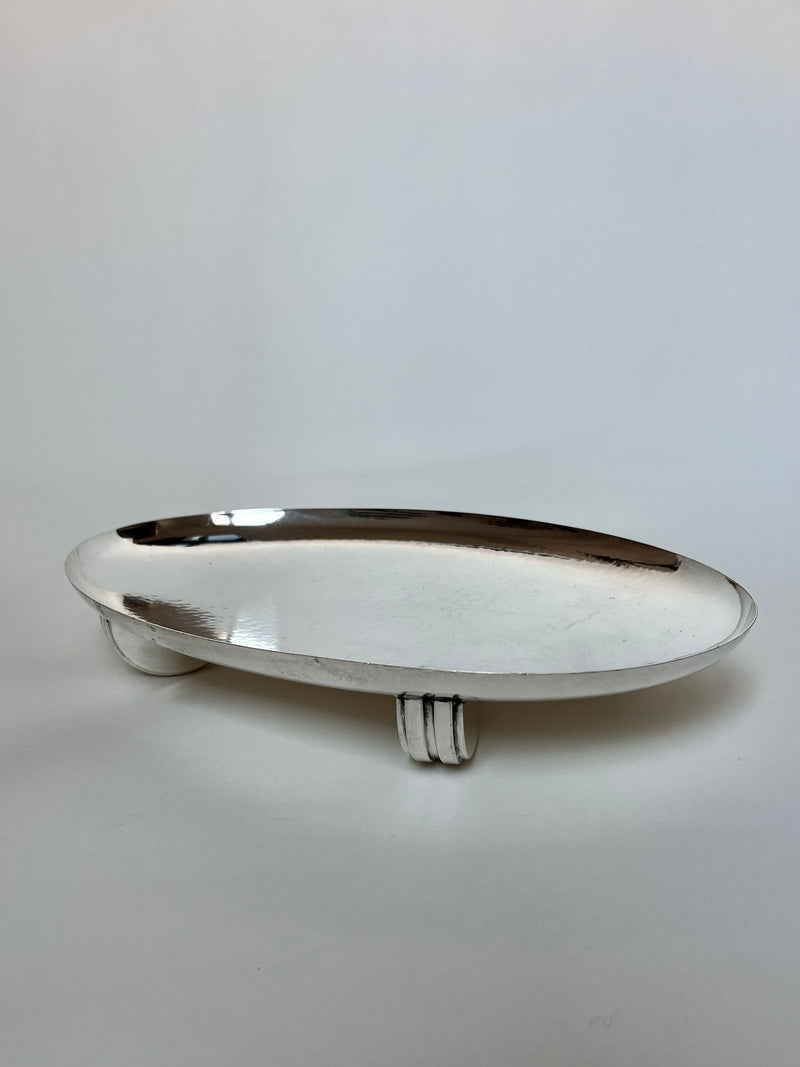 The Manushi Long Art Deco Serving Plate features a sleek, oval reflective surface and stands on three short, curved legs, shown against a plain white background.