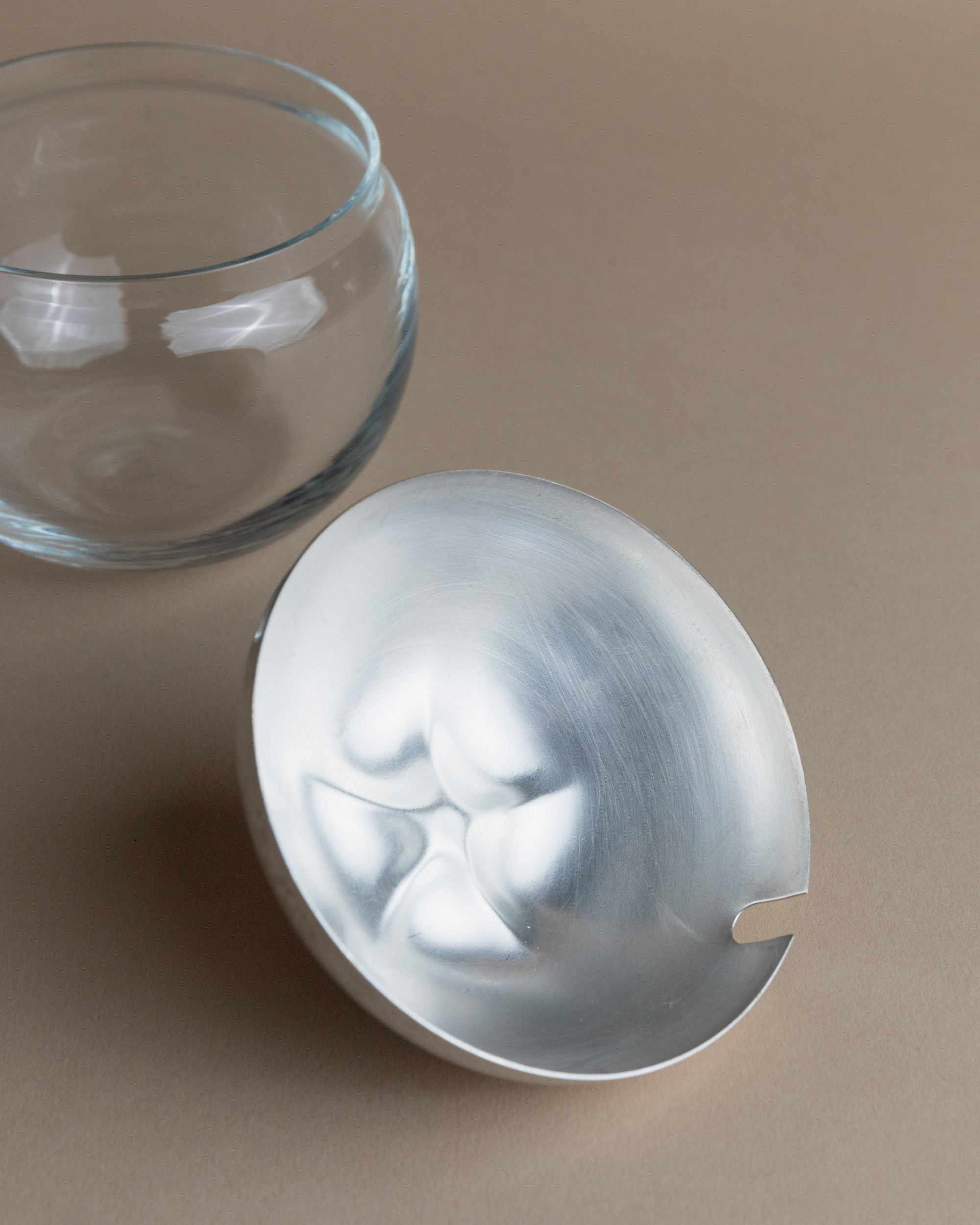 A Bottega Jacobs Silver & Glass Apple Jar sits on a beige surface, accompanied by its shiny, round silver-plated lid with a notched edge. The lid's smooth, reflective surface features a subtle apple-shaped pattern near the center.