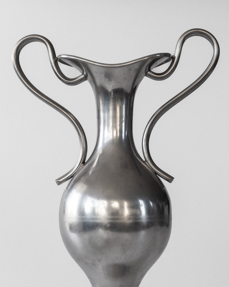 A Pair of Neoclassical Metal Vases by Spigel, Sweden 1953, features a polished silver finish with a smooth, rounded body and elegantly curved handles. Its reflective surface shines brilliantly against a simple light gray backdrop, evoking the elegance reminiscent of Ernst Svedbom's touch.
