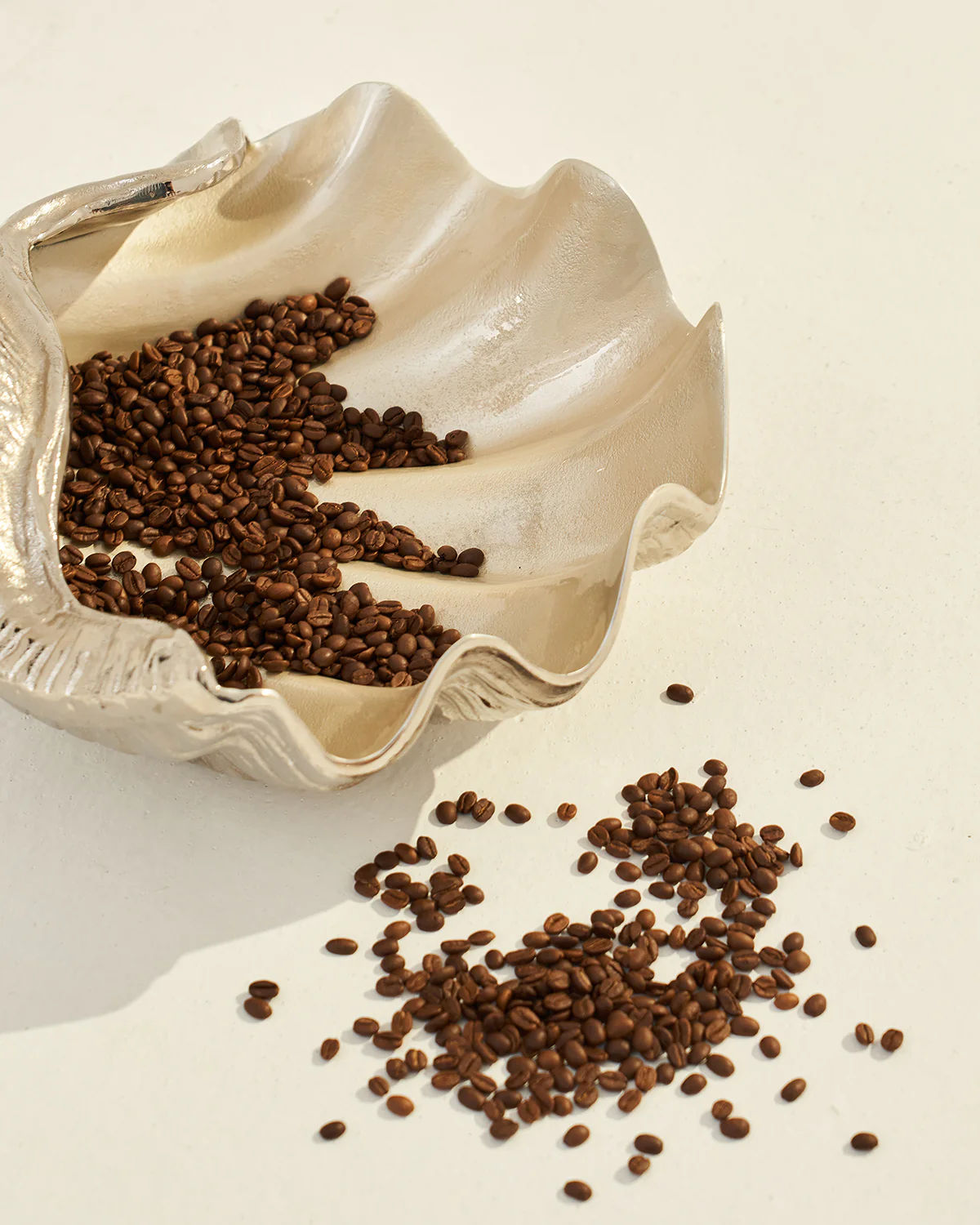 Coffee beans are scattered in and around a silver LMNOH Seashell decorative bowl on a light surface.