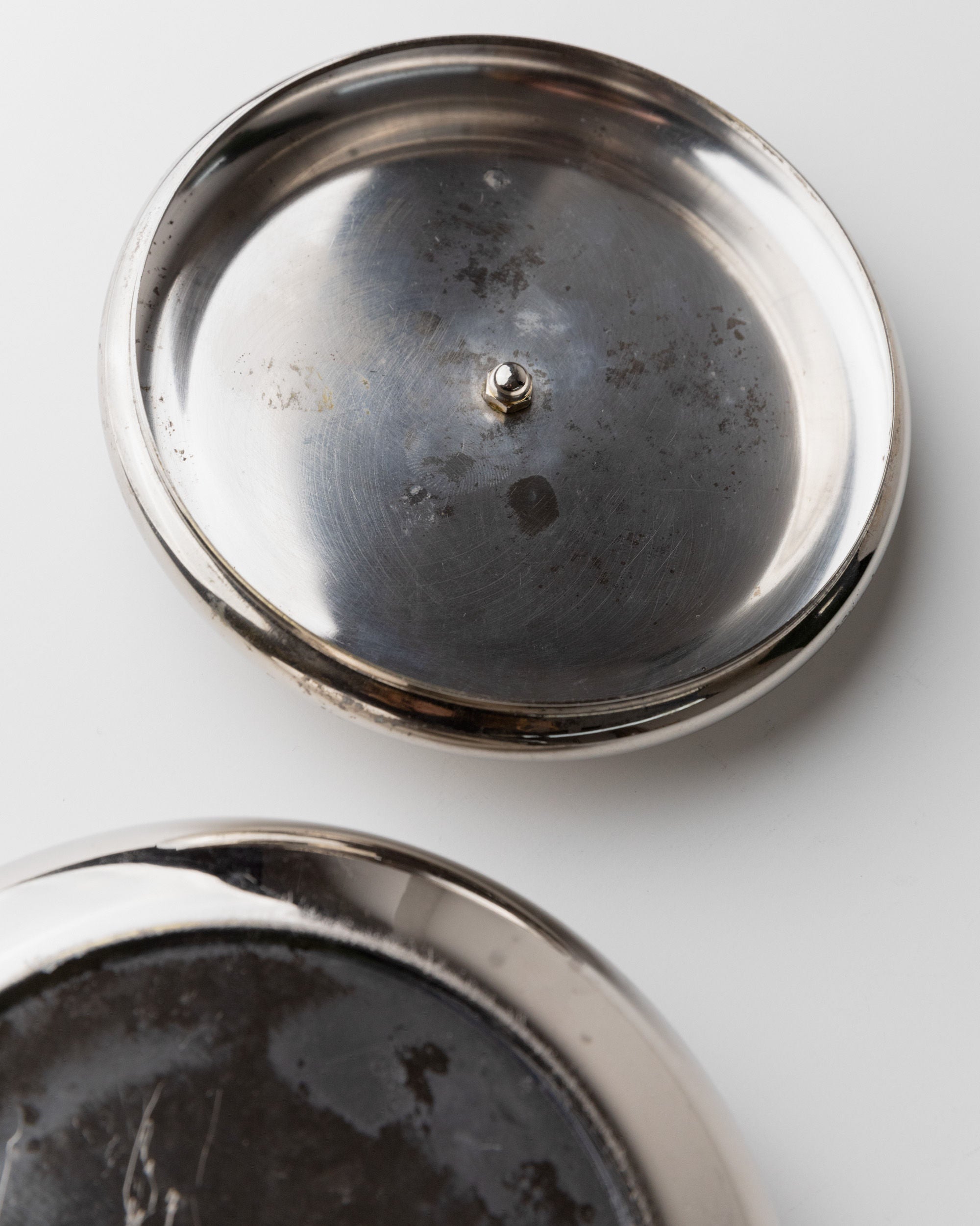 Two vintage Bottega Jacobs silver-plated box lids from 1950s Italy rest on a white surface. The foreground lid is partially visible, and the background lid features an acorn handle. Both exude a tarnished, nostalgic charm.