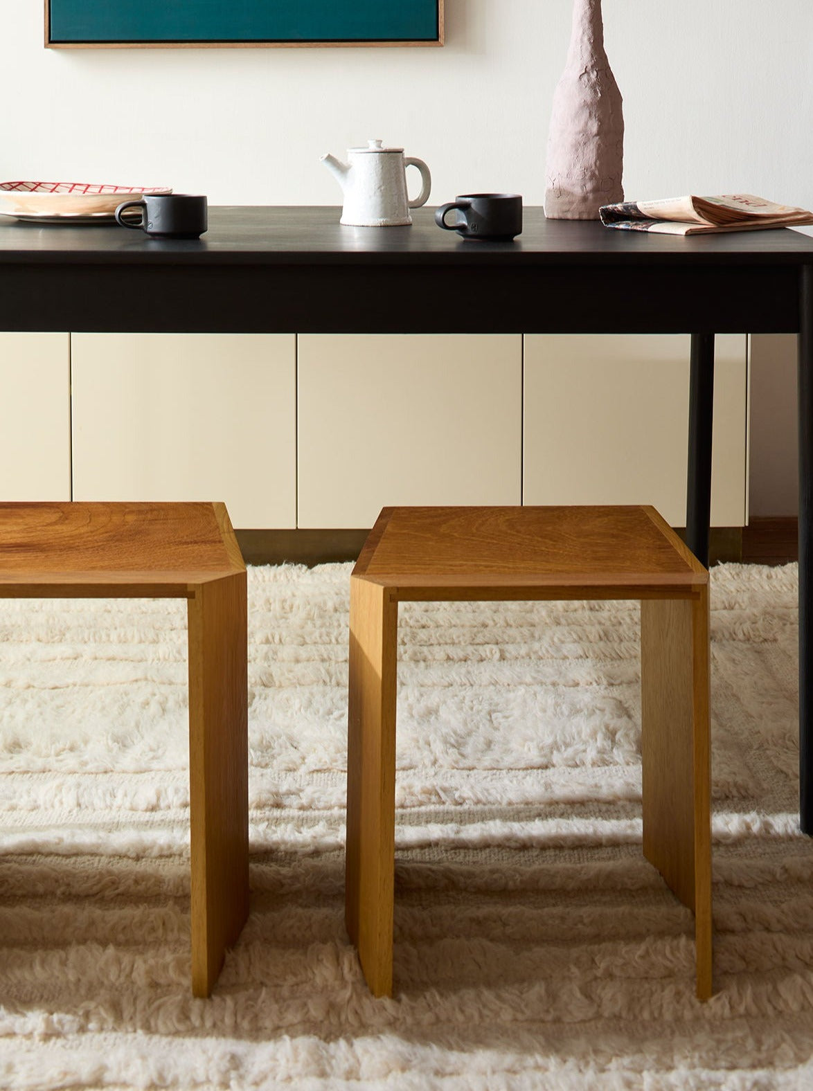 A modern dining area featuring a sleek black table with two Lemon Athens Stools in Iroko, set on a plush white rug. Accessories include a white kettle and beige ceramics.