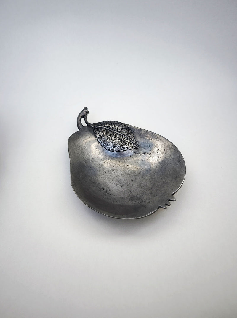 The Les Objoies Pear Vide-poches is a small, pear-shaped dish with a raised leaf design near the top and an antique brass finish, shown against a plain white background.