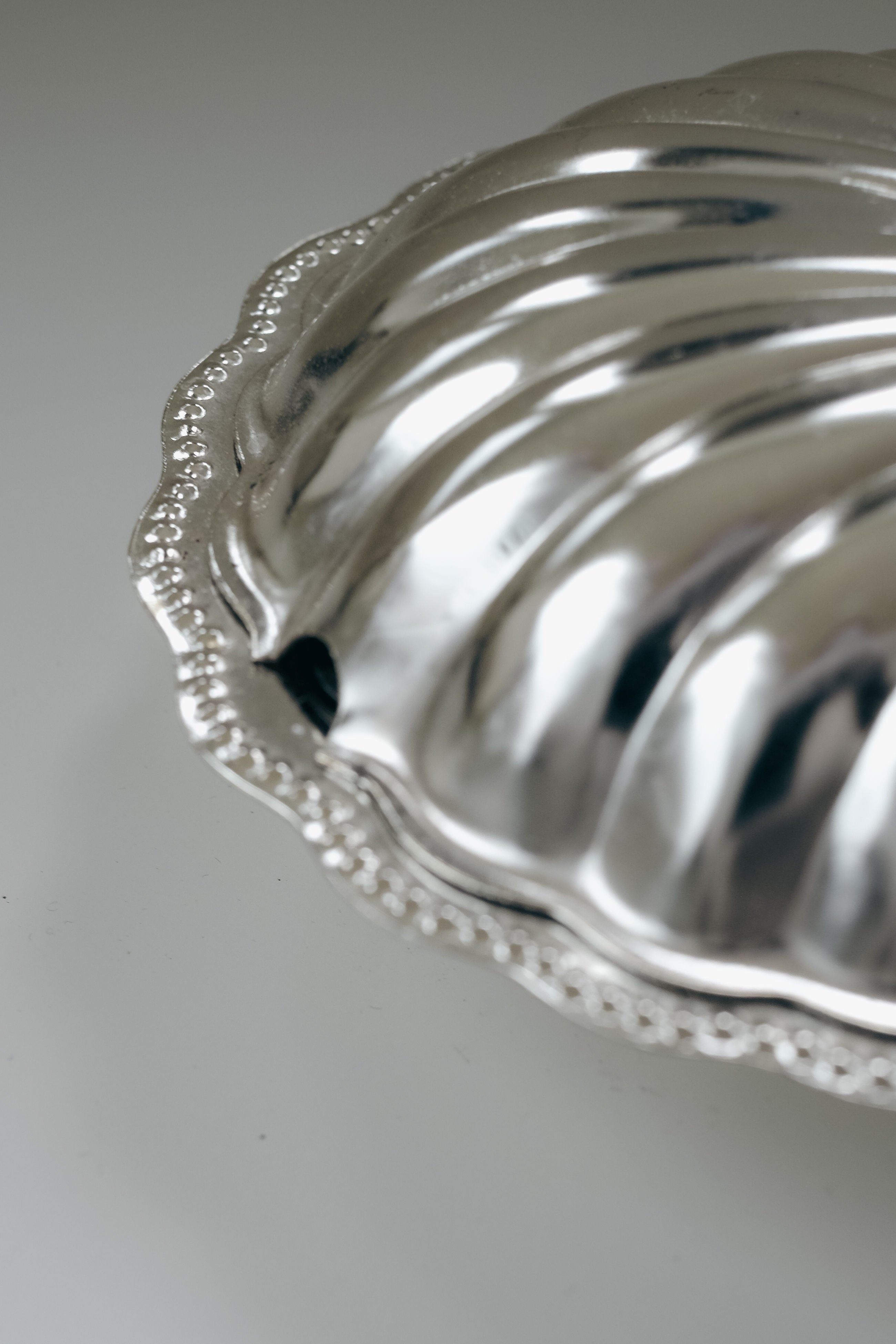 Close-up of a septembre studios Shell Shaped Butter Dish featuring a textured, scalloped silver edge and shiny reflective surface with swirling patterns, set against a soft, neutral background.