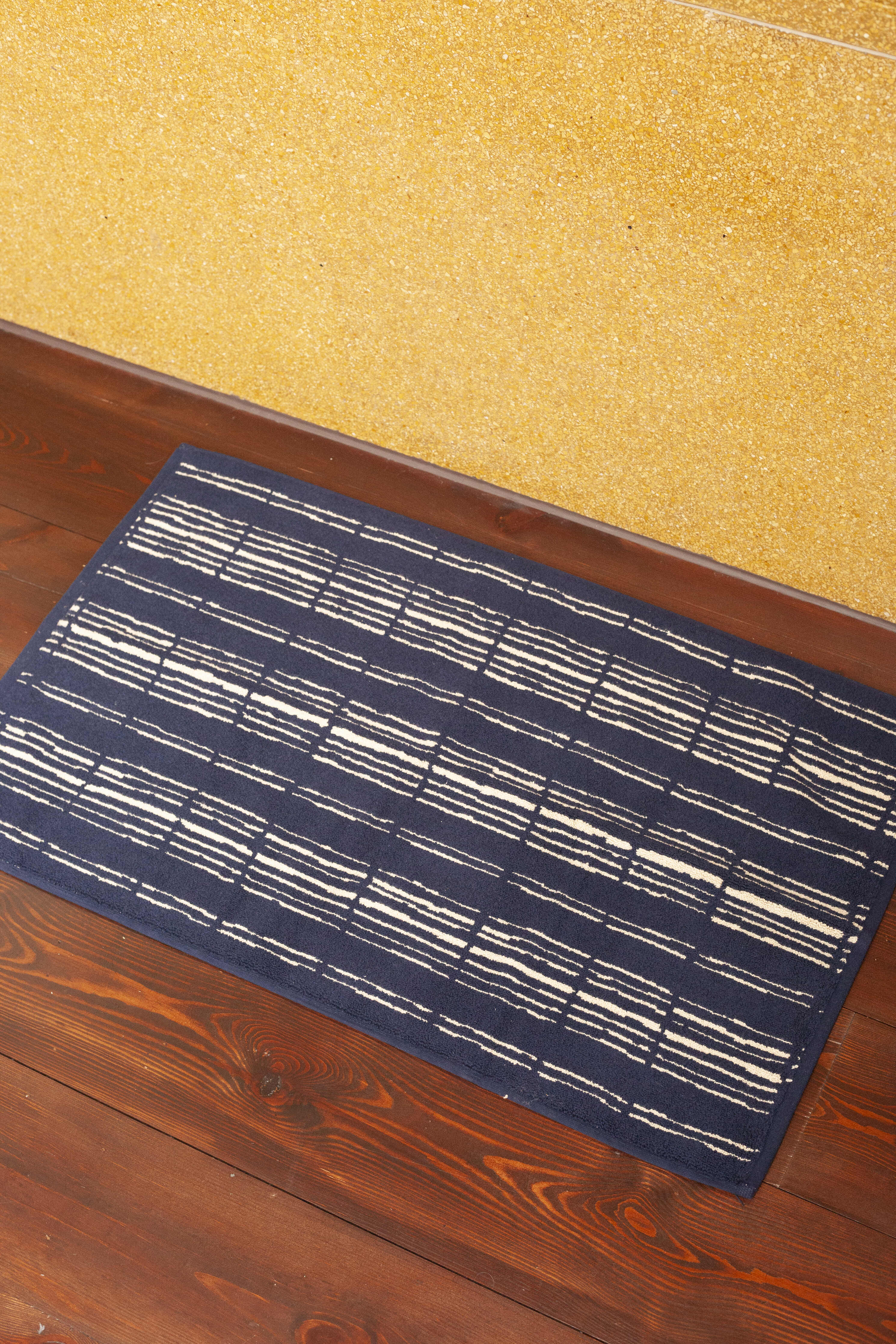 A rectangular dark blue Autumn Sonata Ester Bath Mat with a white linear pattern is placed on a wooden floor, adjacent to a textured yellow wall. The design on this 100% cotton mat consists of repeating horizontal and vertical lines, creating a grid-like appearance that exudes luxurious comfort.