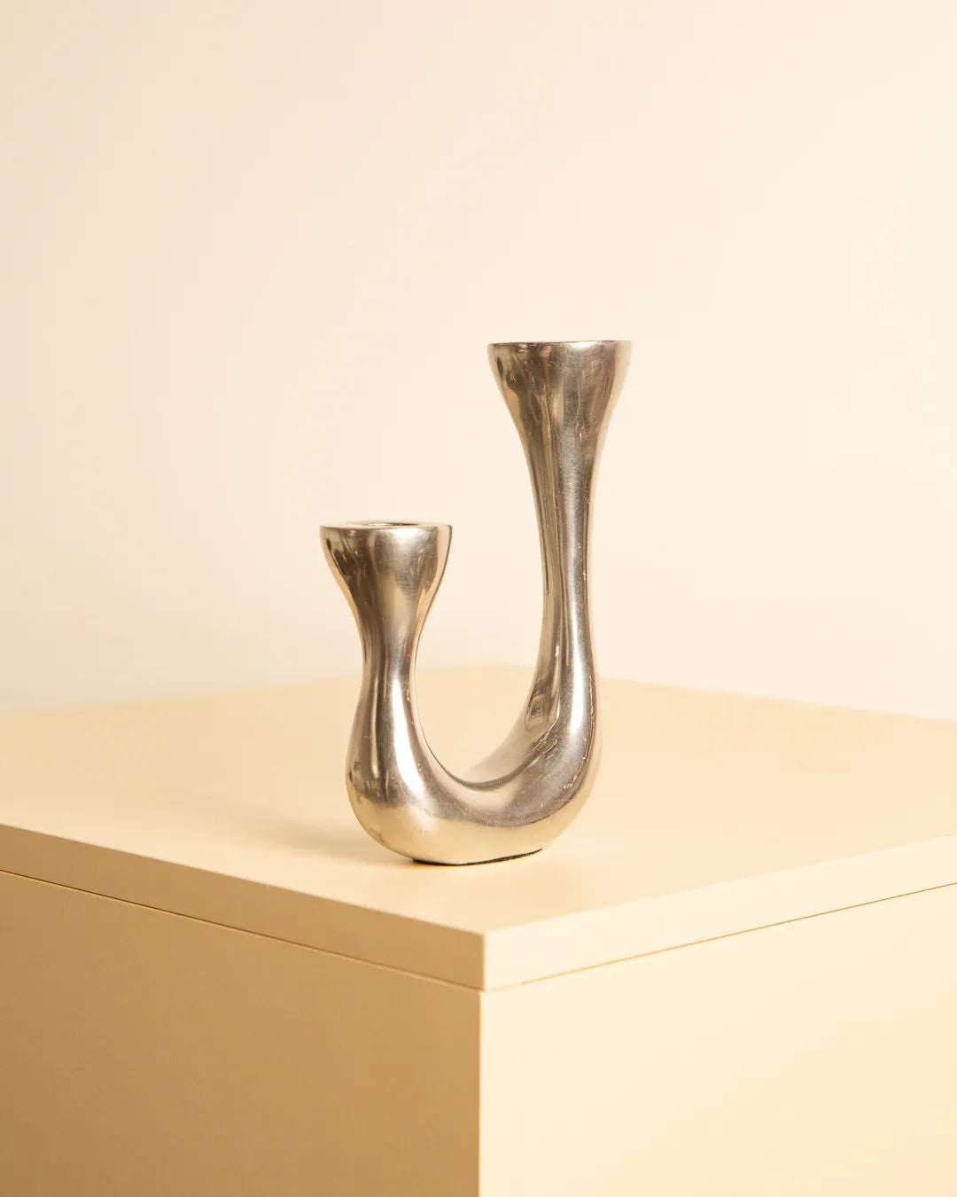 Treaptyque's 80s Biomorphic Candleholders feature a sleek, avant-garde design with two asymmetrical arms of differing heights. Resting on a light beige surface against a matching beige background, this modern silver piece boasts smooth, curved lines that enhance its minimalist aesthetic.