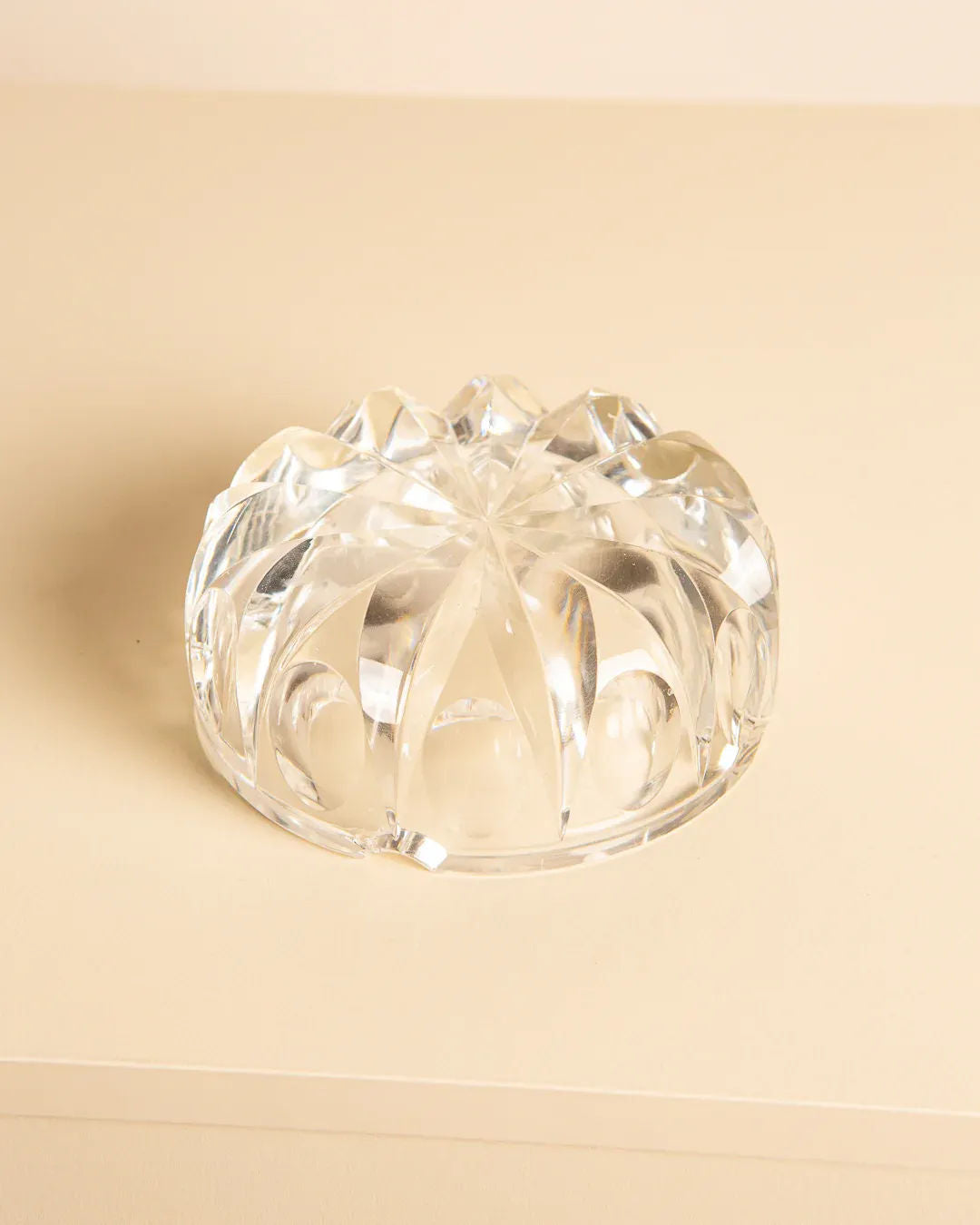 A round, clear crystal Saint-Louis “Ambassadeur” D120 ashtray from the 1970s by Treaptyque sits on a beige surface, featuring intricate faceted patterns and geometric shapes that reflect light.