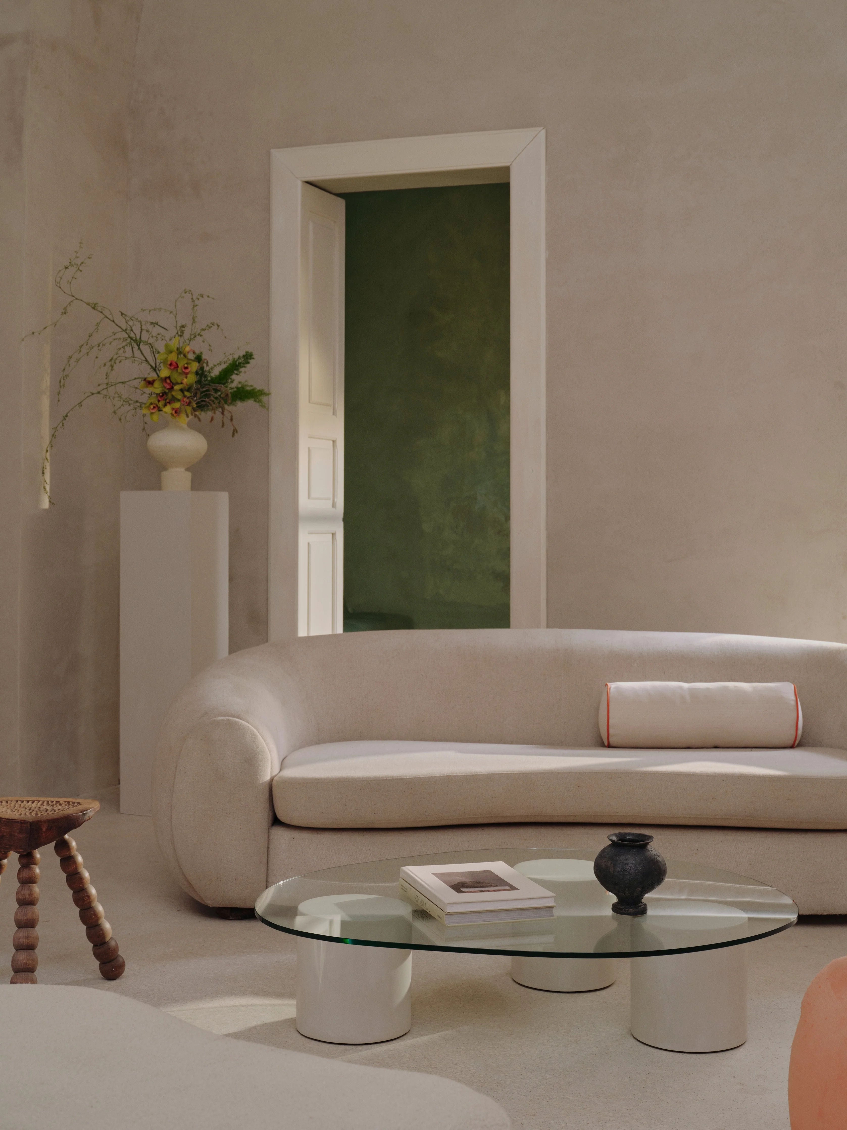 A minimalist room showcases a curved beige sofa accompanied by the "Mattia" Glass Coffee Table from MarlotBaus, featuring a tempered glass top that supports a book and a black vase. A tall white pedestal highlights a flower arrangement, while an open door reveals a green wall in the background.