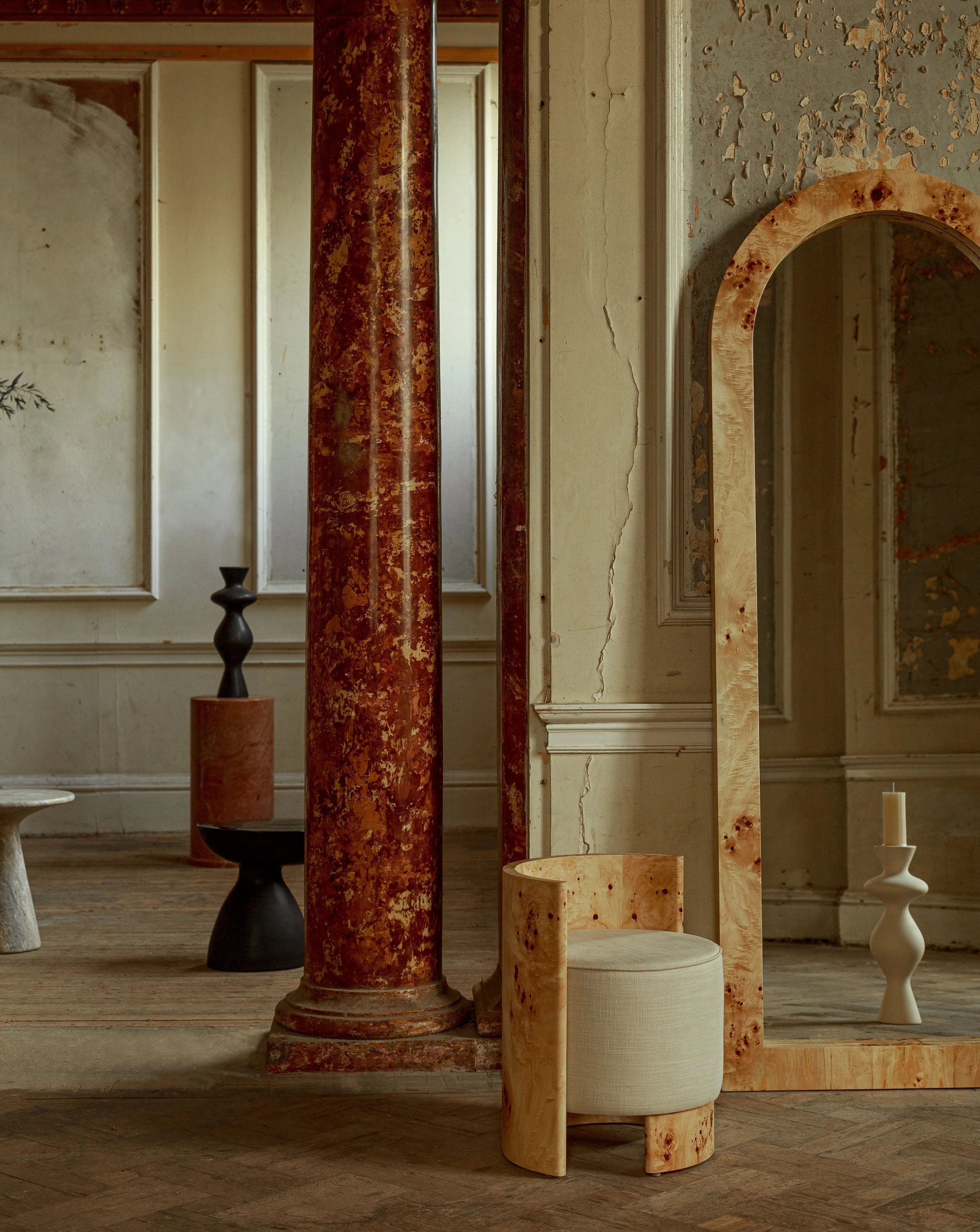 A rustic interior showcases a tall arched mirror next to the minimalist "Livon" Burl Armchair by Ransom & Dunn. Nearby stands a weathered red marble column, while a black sculptural vase rests on a small table. The textured, aged walls enhance the vintage ambiance with subtle touches of ash burl wood details.