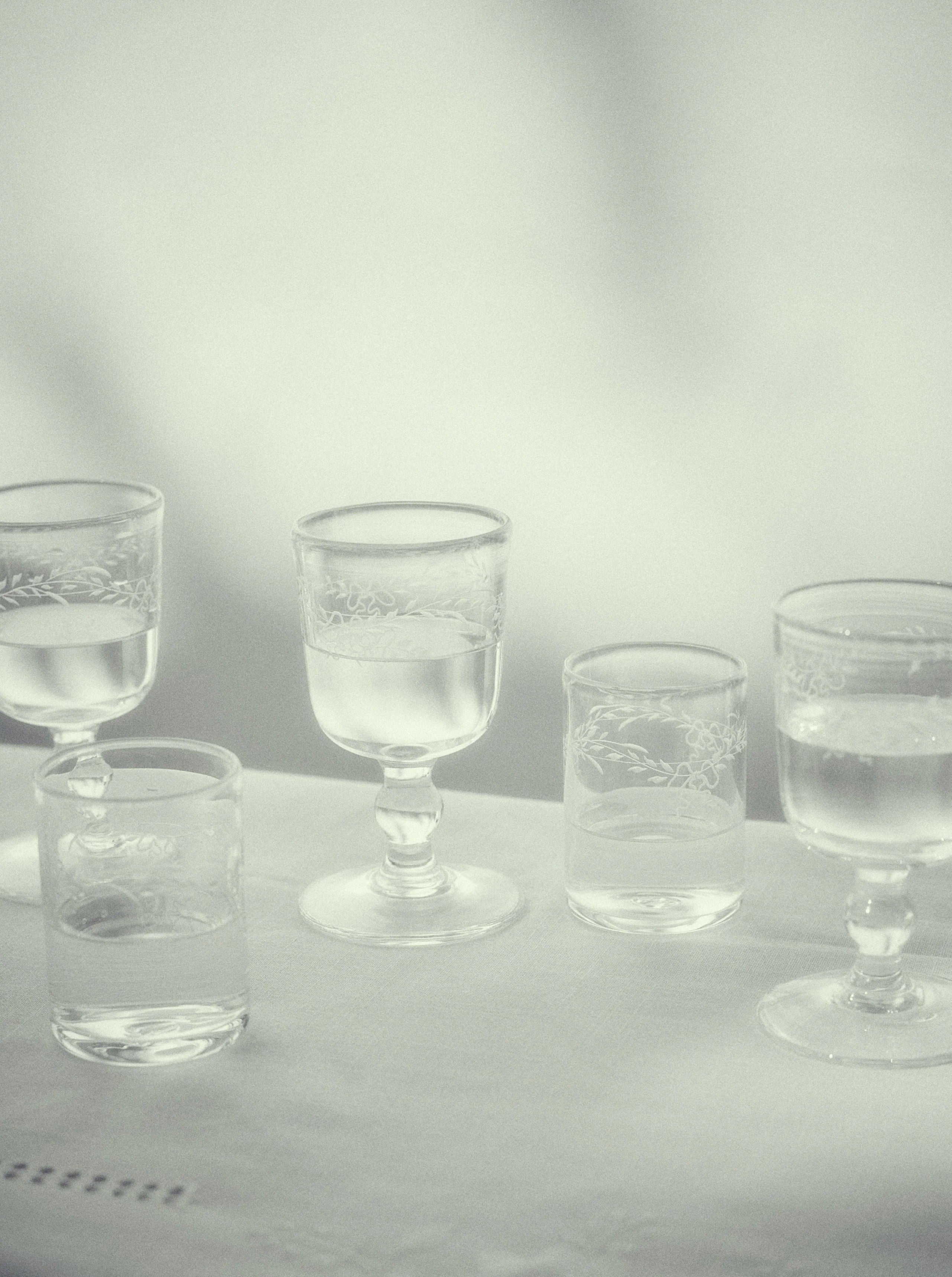 Monochrome image of various Akua Objects Barbro Water Glasses, set on a table with a textured tablecloth. The background has soft, blurred shadows, creating a serene ambiance.