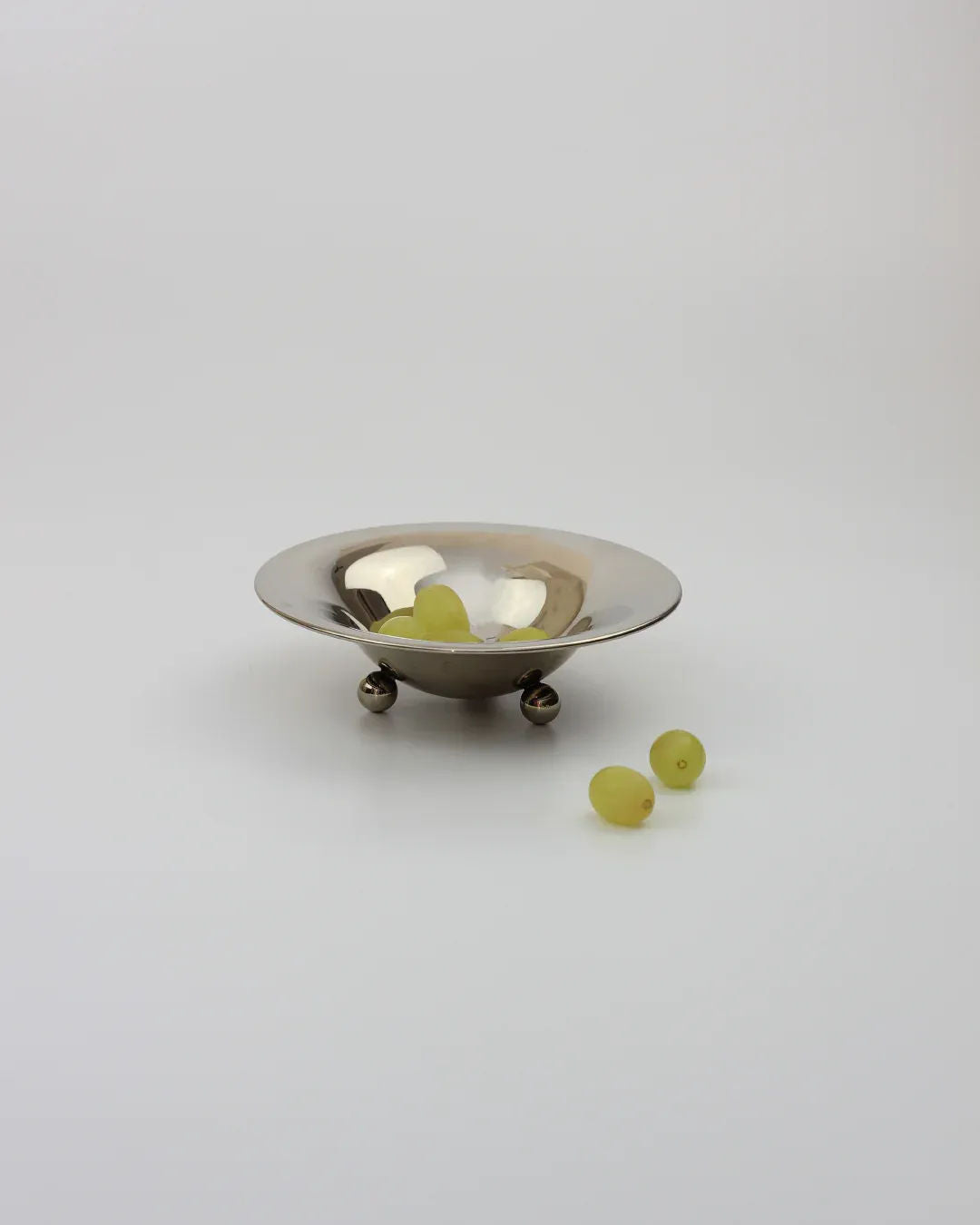 A small silver "Bowl on Three Legs" by Boga Avante Shop, holding a handful of green grapes, sits on a plain white surface with two grapes outside casting shadows. It reflects the simplistic elegance of late 20th-century design.