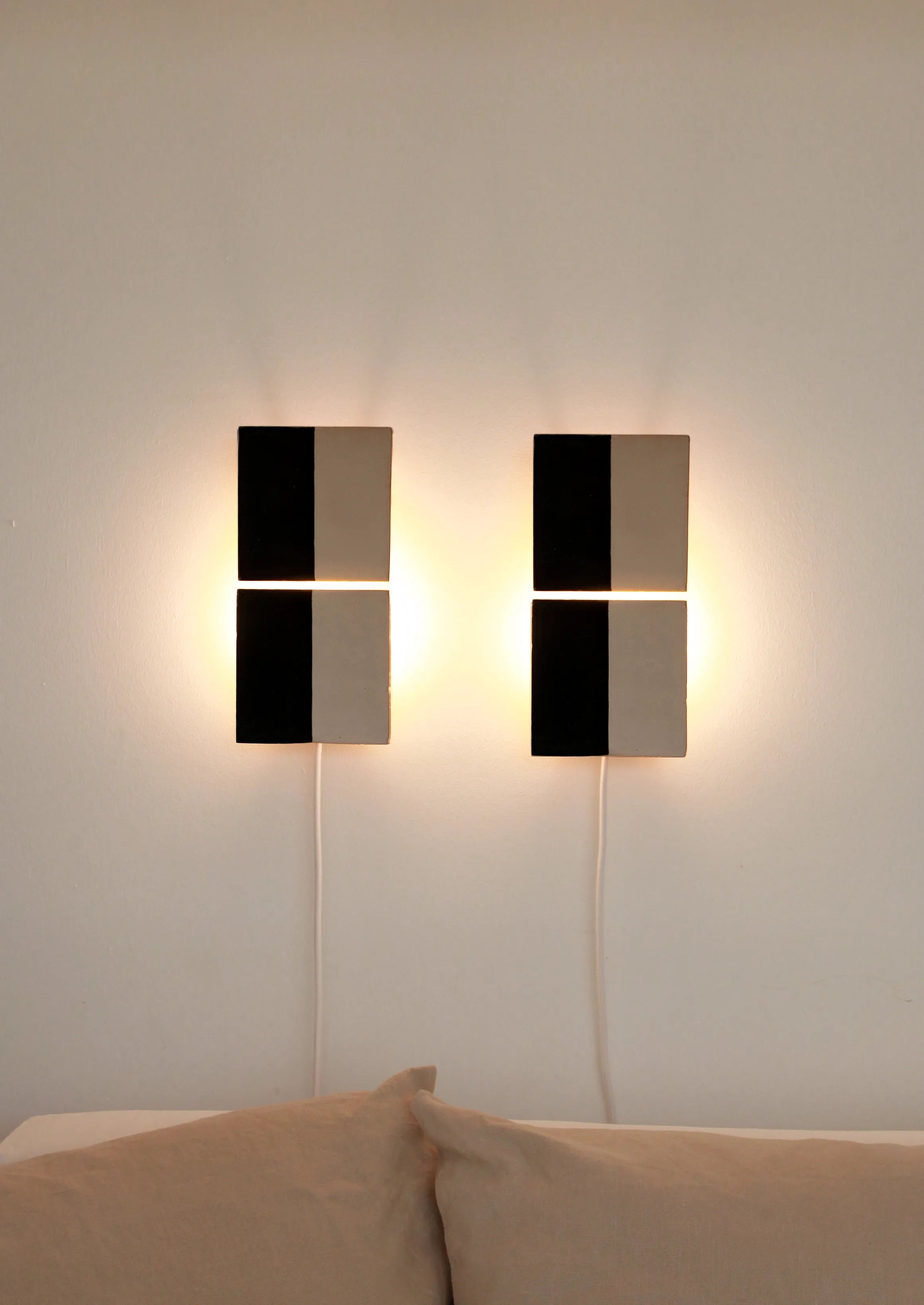 Two Tiles Line N ceramic wall lights from Violaine d'Harcourt, featuring geometric black and white halves, are mounted side by side on a white wall. A soft glow illuminates the bed below, adorned with beige pillows.