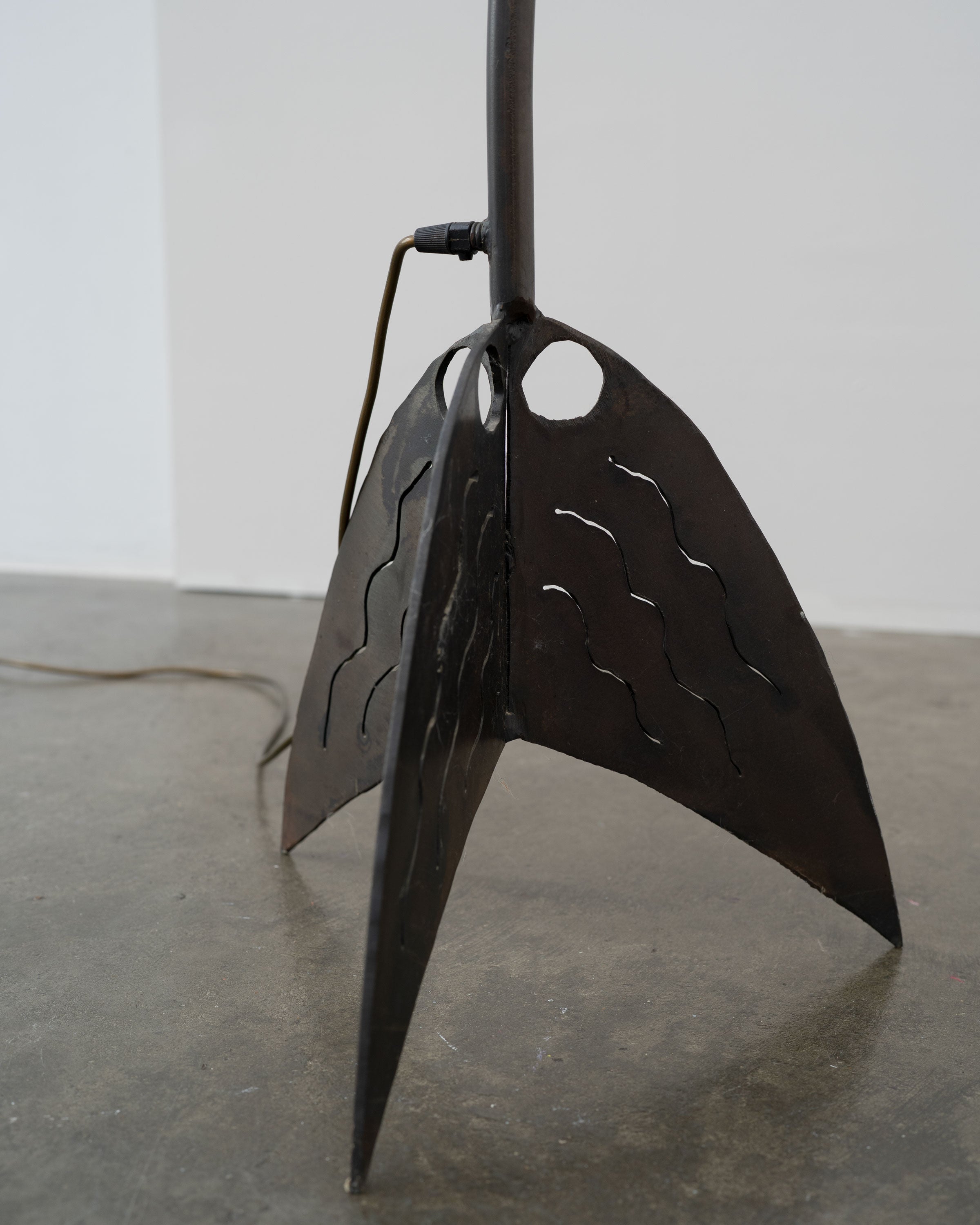 A close-up of the Brutalist Wrought Iron and Cast Glass Lamp by Tom Bogle, Norway, c.1970, with three curved, wavy-cutout legs and a visible cord, standing on a concrete floor against a plain wall.