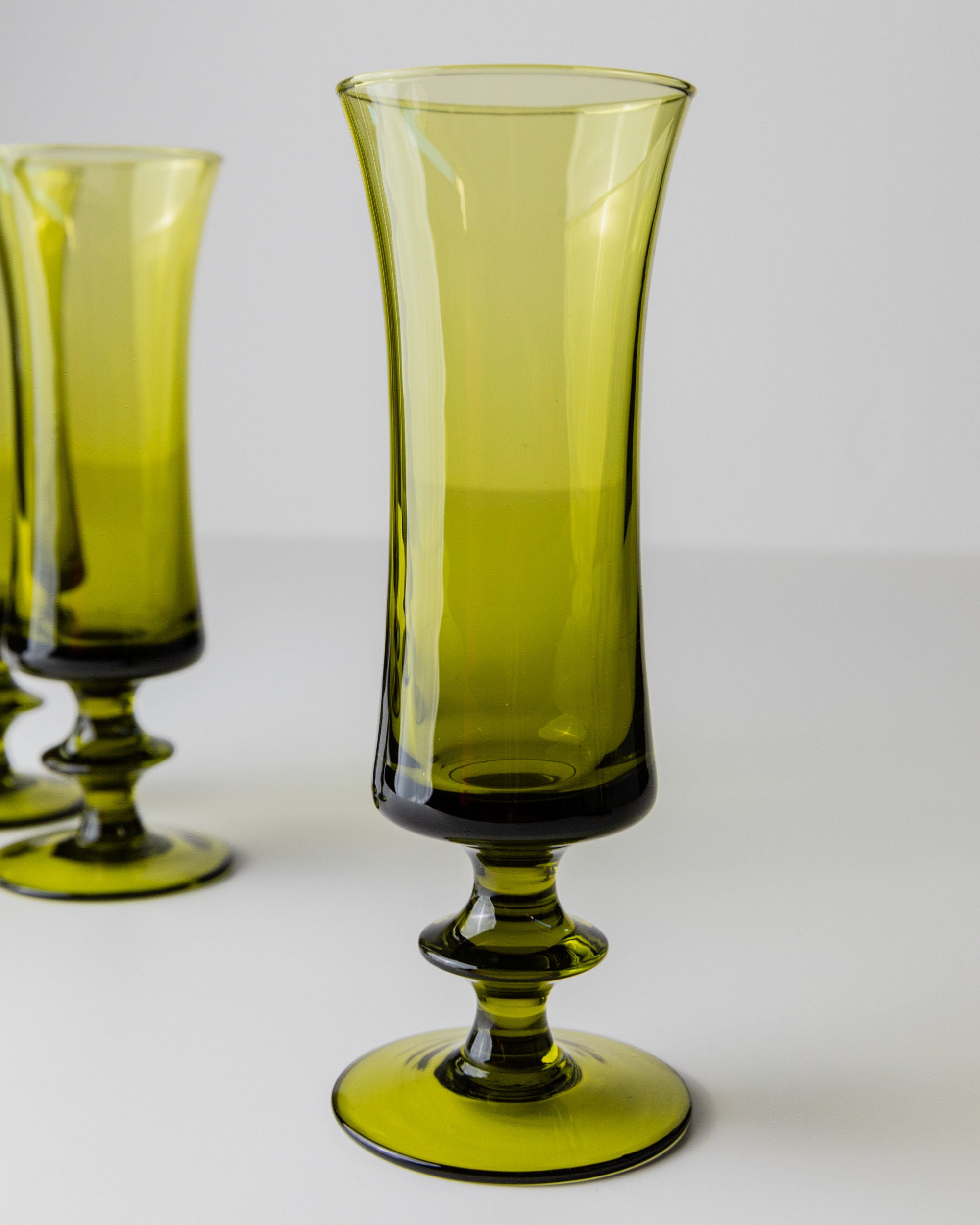 The white surface showcases Bottega Jacobs' 4 Murano Glasses in Green 70s, featuring a tall, elegant design and wide bases. One goblet is in focus, evoking vintage charm, while two blurred goblets in the background add a retro chic flair.