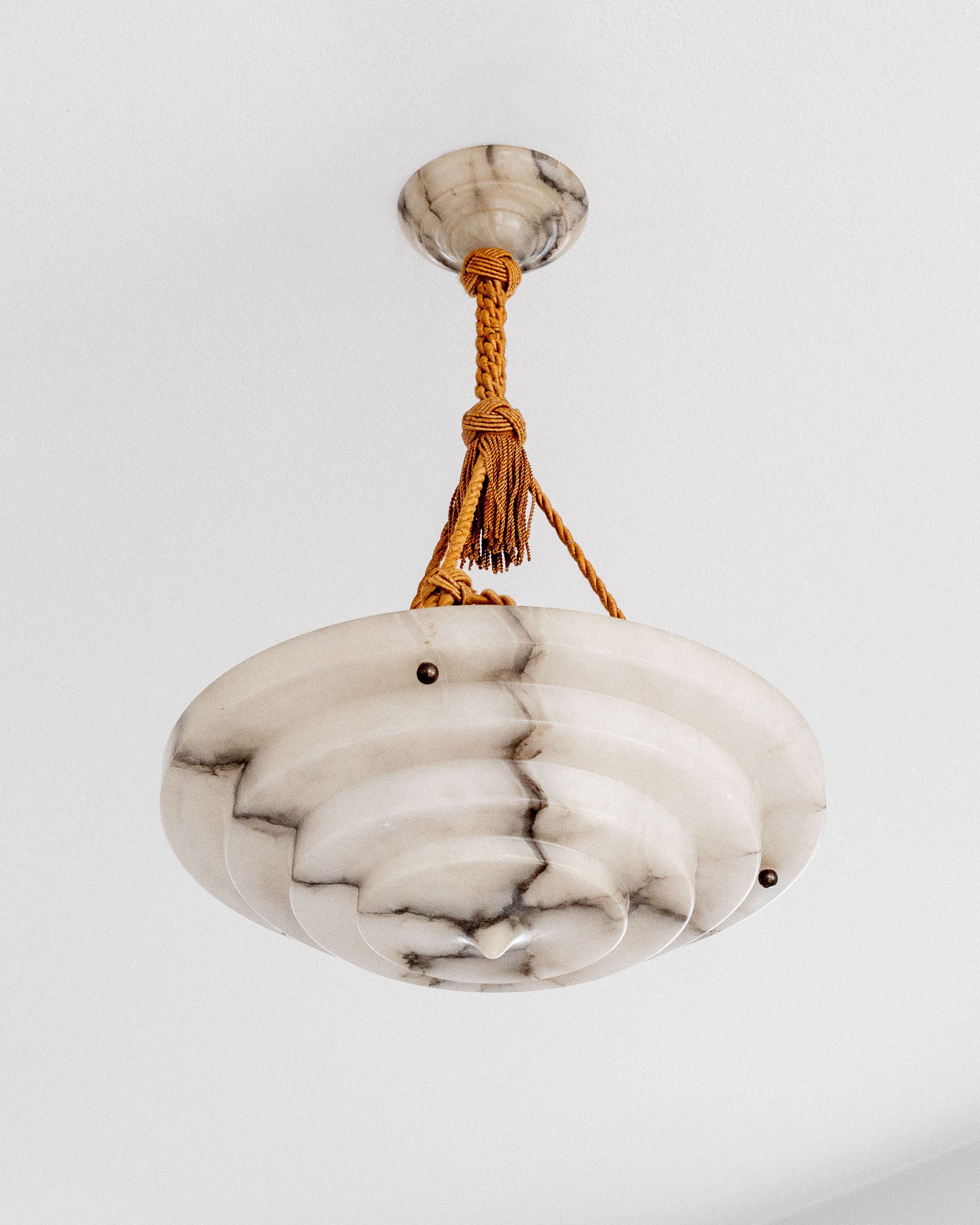 The Spigel Art Deco Alabaster Pendant Chandelier is a stylish fixture with a round, tiered design resembling marble or stone. It boasts black vein patterns and is suspended by a decorative braided orange cord.