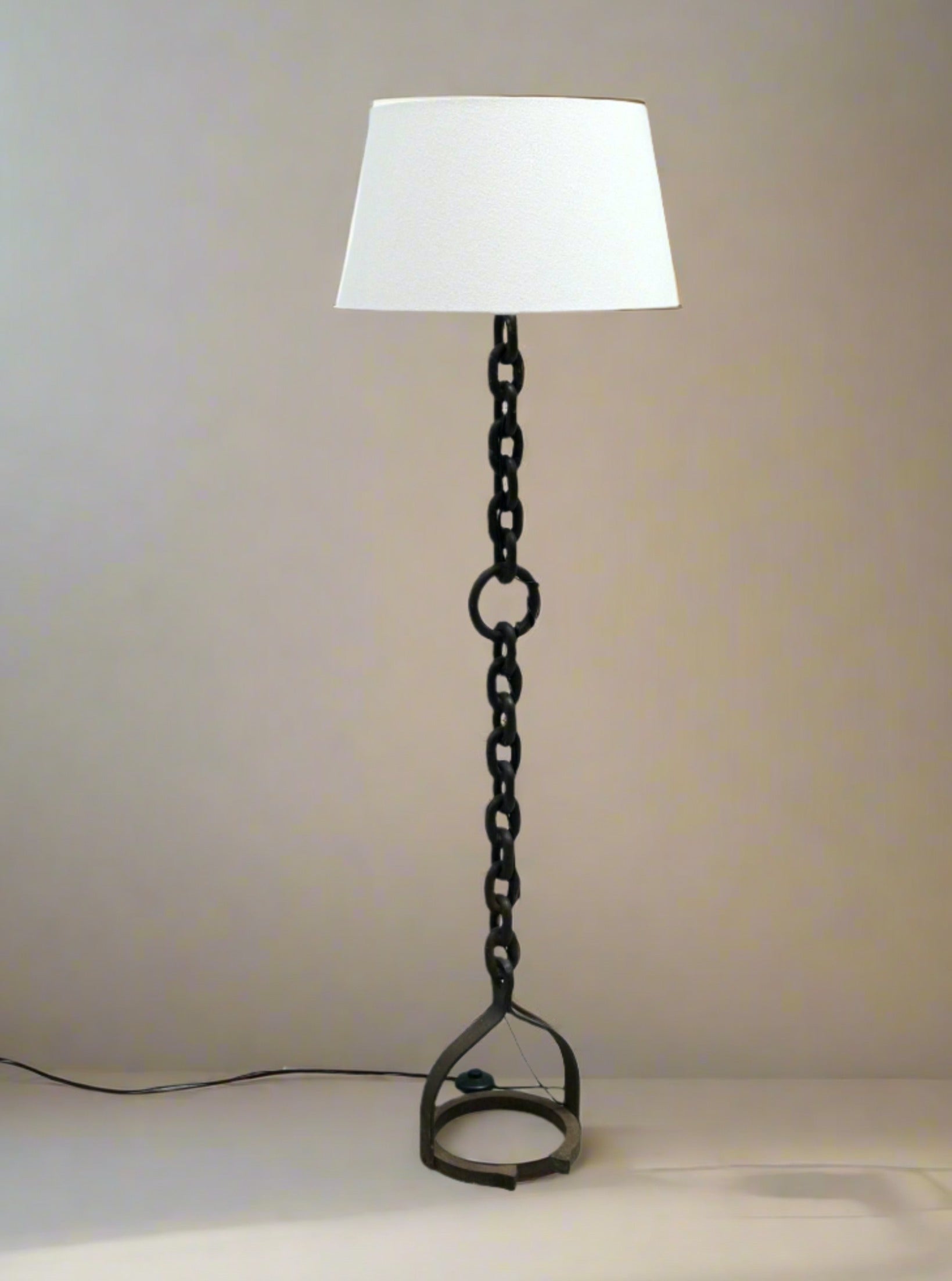 This distinctive Lampadaire chaîne brutaliste by LA NOUVELLE GALERIE boasts a white shade paired with a base that mimics iron chains and a stirrup. Set against a simple beige wall, it gracefully rests on a light surface, exuding subtle style de Ryckaert influences.