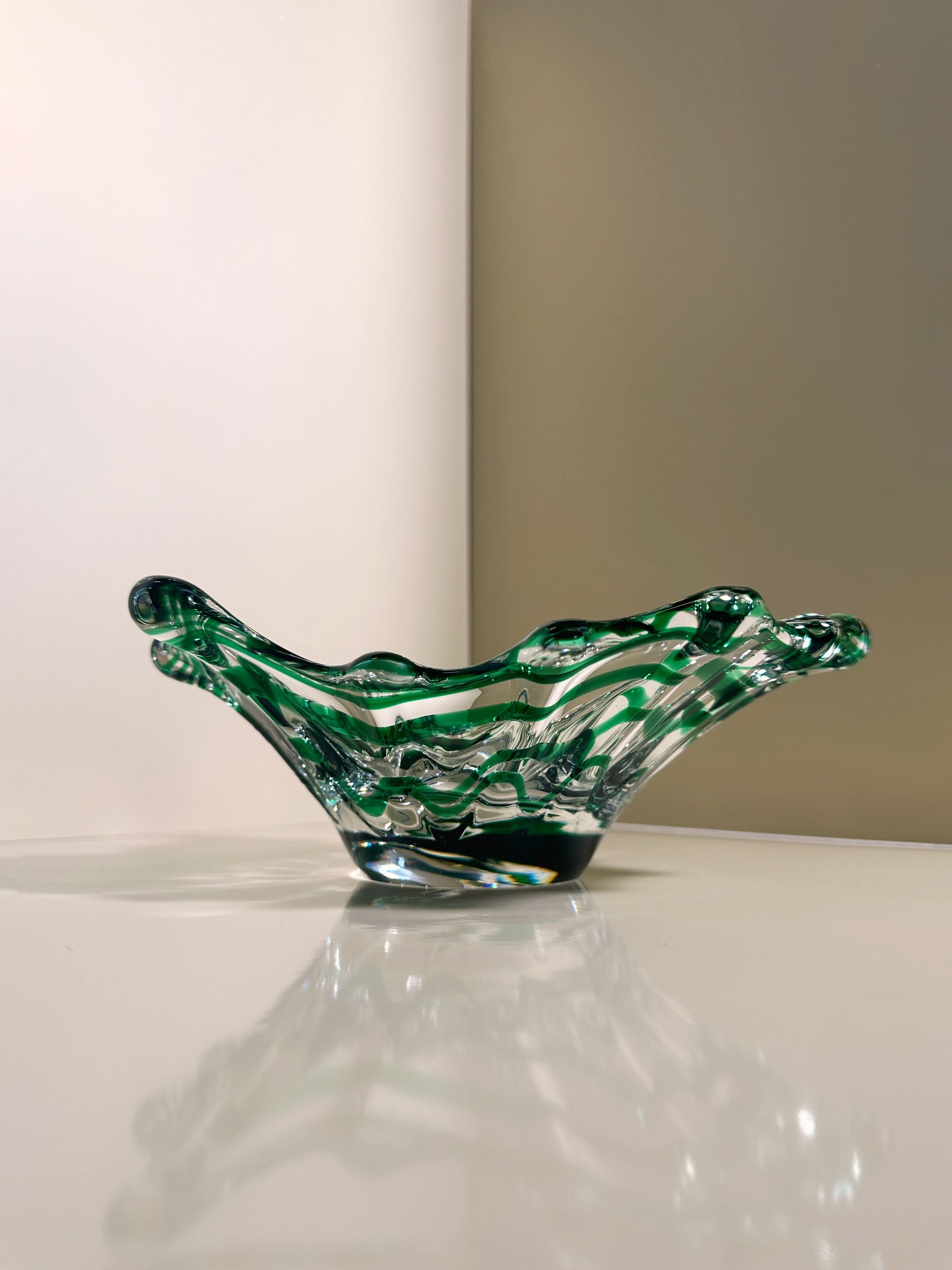 The Green Swirled Murano Vide-Poche by Dodo Vintage, with its wavy, artistic edges and intricate swirling pattern, rests elegantly on a reflective white surface against a beige backdrop, casting subtle reflections reminiscent of classic Murano artistry.