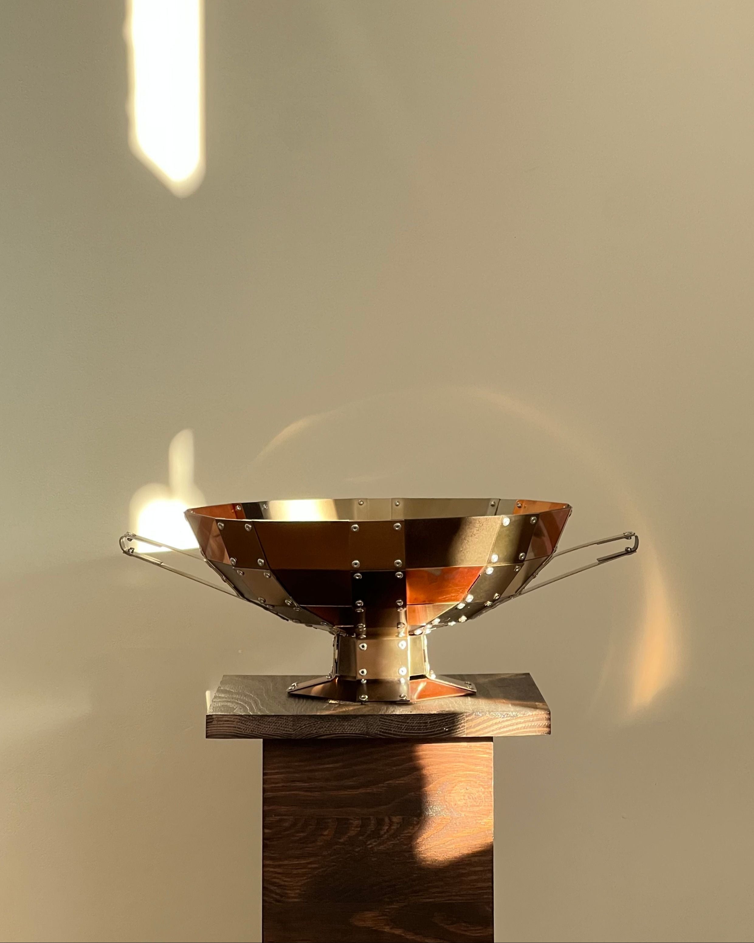 The Kylix by Theo Galliakis, a metallic riveted bowl inspired by Ancient Greece, rests on a dark wooden pedestal. Warm sunlight and soft shadows highlight its industrial finish against a minimalist beige wall.