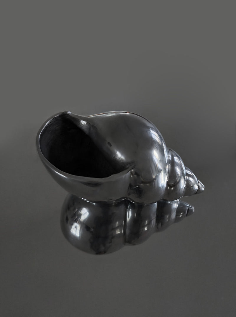 The Large Shell Sculpture by Les Objoies is displayed on a reflective surface, its glossy black spiral design mirrored below. A solid dark gray background enhances the sculpture’s lustrous texture.