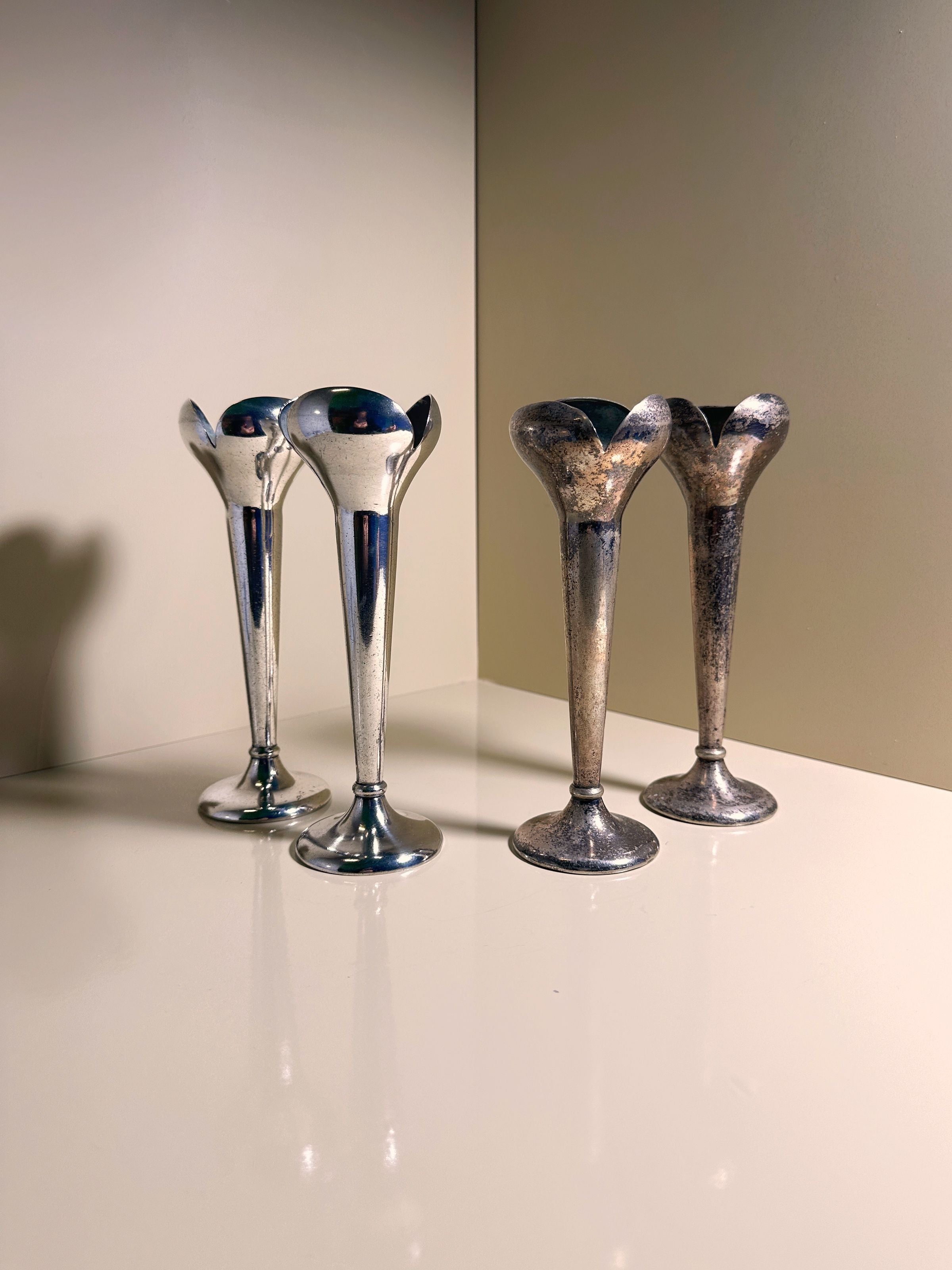 Two Alpacca silver vases with a polished shine reflect the elegance of the 1930s Gio Ponti candlesticks, while the other two vases display a tarnished charm on a beige surface against a neutral wall.