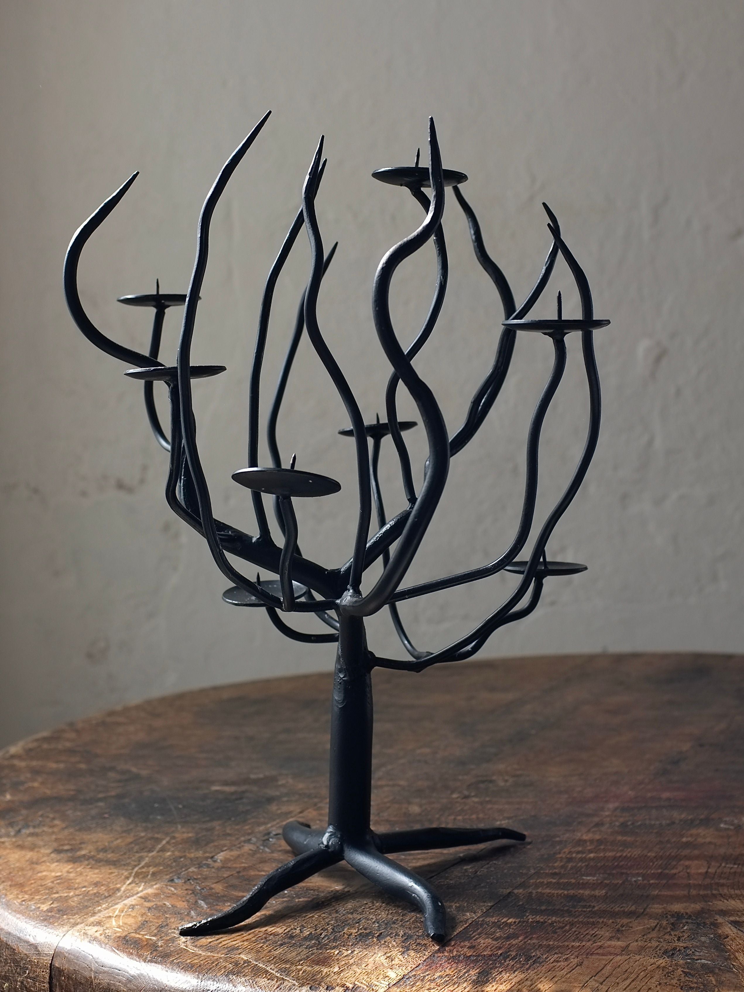 The Hand-forged Metal Sculptural Candle Holder from Veter Vintage, with its black metal finish and 1960s-inspired design, stands proudly on a rustic wooden surface. Its multiple branches are expertly crafted to hold small candles, creating a striking silhouette against the plain, light-colored wall.