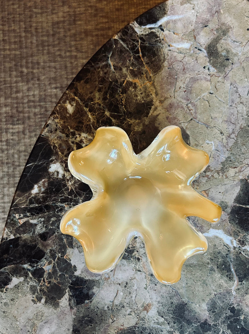 A Murano Glass Dish by Flock Studio, shaped like a flower and featuring a vibrant yellow color, rests on a marbled surface. The lighting accentuates its smooth edges and glossy texture, making this exquisite piece of art an embodiment of elegance and craftsmanship.