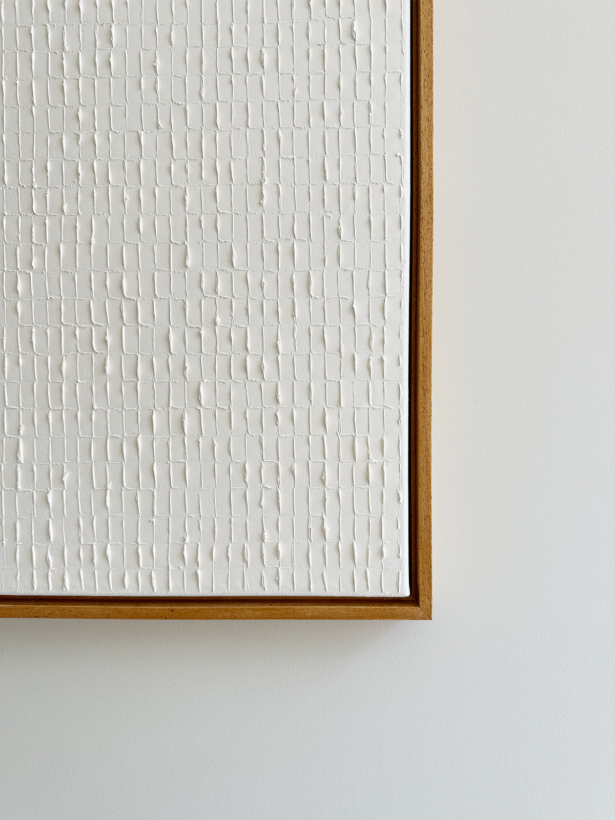 A close-up photograph of an abstract piece of art inspired by the Korean Dansaekhwa movement. The artwork features a textured, white grid pattern on a white background, evoking mindfulness. It's framed in a simple wooden frame and hung against a light-colored wall, with minimal and uniform texture. The piece is titled "Warm White - Oil on canvas" by Roi Elmaliah.