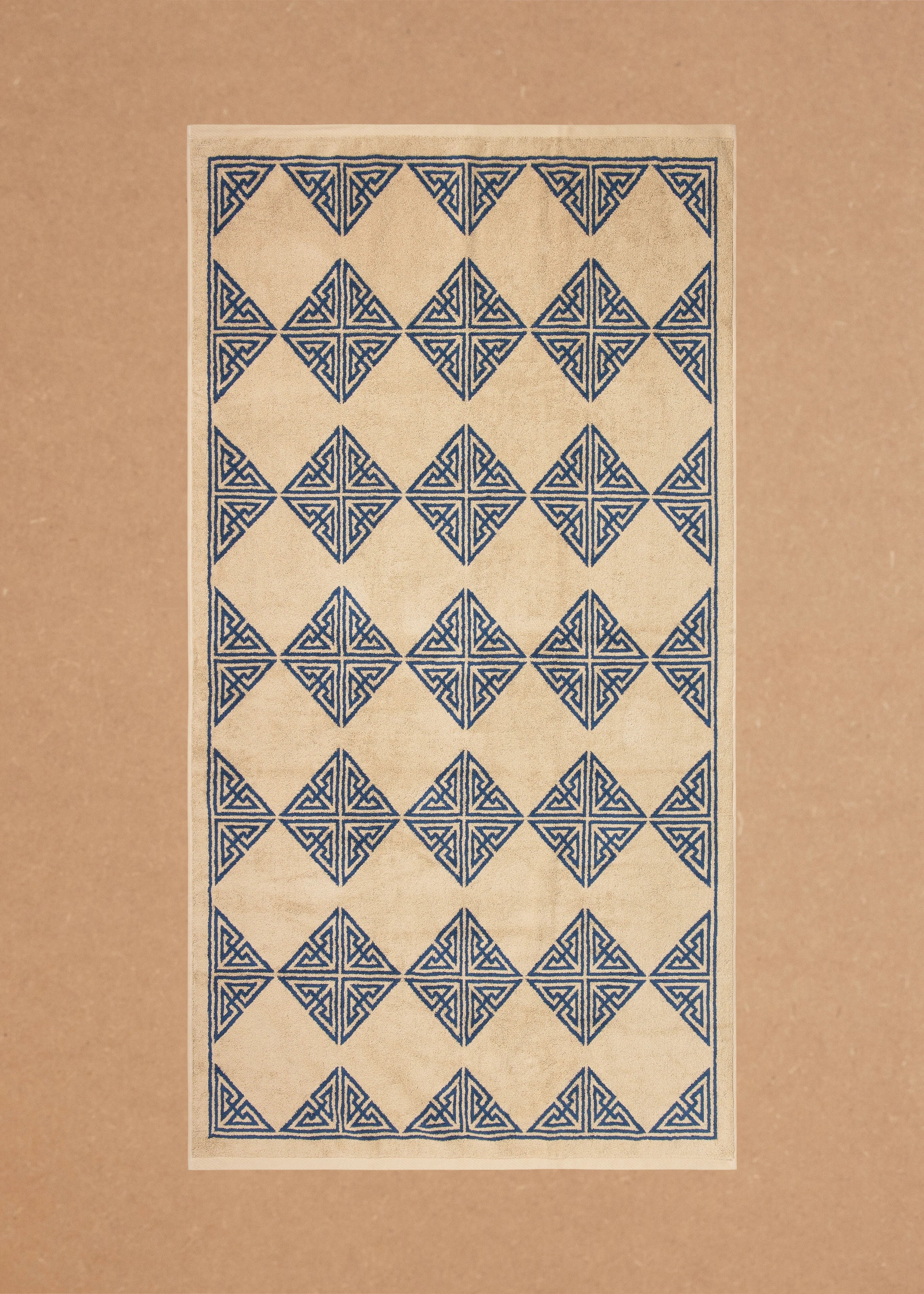 Autumn Sonata Agnes Bath Towel featuring a symmetrical geometric pattern with blue triangles and diamonds on a beige background, presented on a tan surface, made in Portugal.