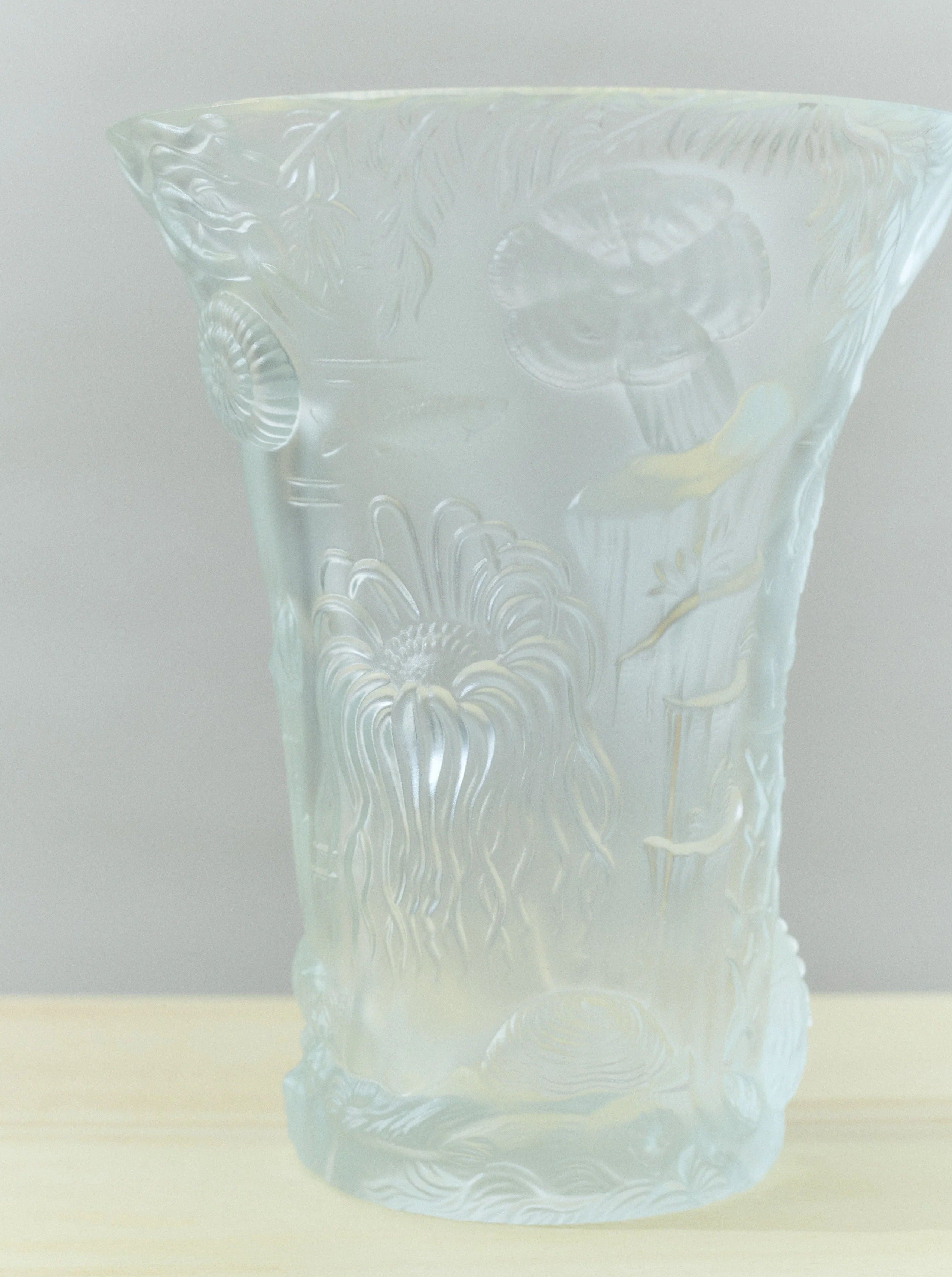 A handmade Michael Lagoon Vase by Akua Objects with intricate etched floral and leaf designs sits on a light wooden surface. The translucent material showcases delicate details, creating a soft, elegant appearance. Reminiscent of Bohemian Glassware, the vase's top flares outward subtly.