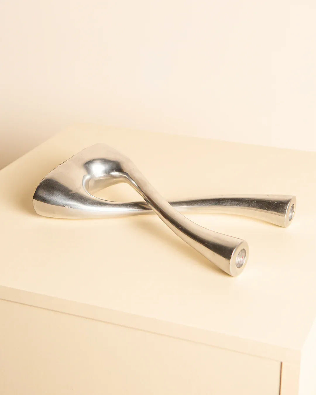 The Aluminum “Croisé” Candleholder 80's by Treaptyque is a sleek, modern metallic sculpture with a wishbone shape that radiates timeless allure. Placed on a light-colored surface against a neutral background, its unique design captivates attention.