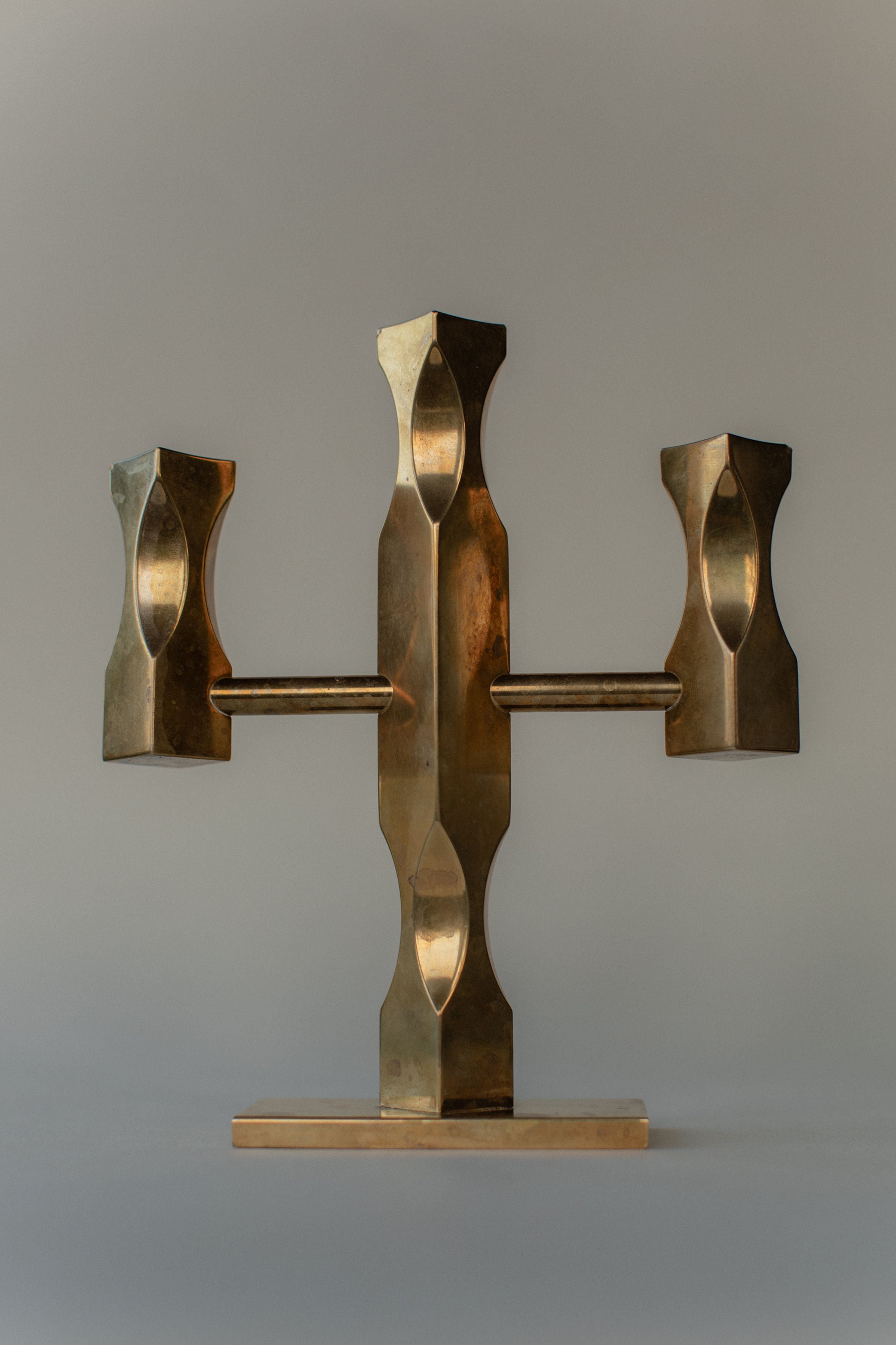 The Brass Candleholder by Out For Lunch is a bronze sculpture with a geometric design, symmetrical shapes, and square elements. It resembles a Brutalist style and stands on a rectangular base against a neutral background.