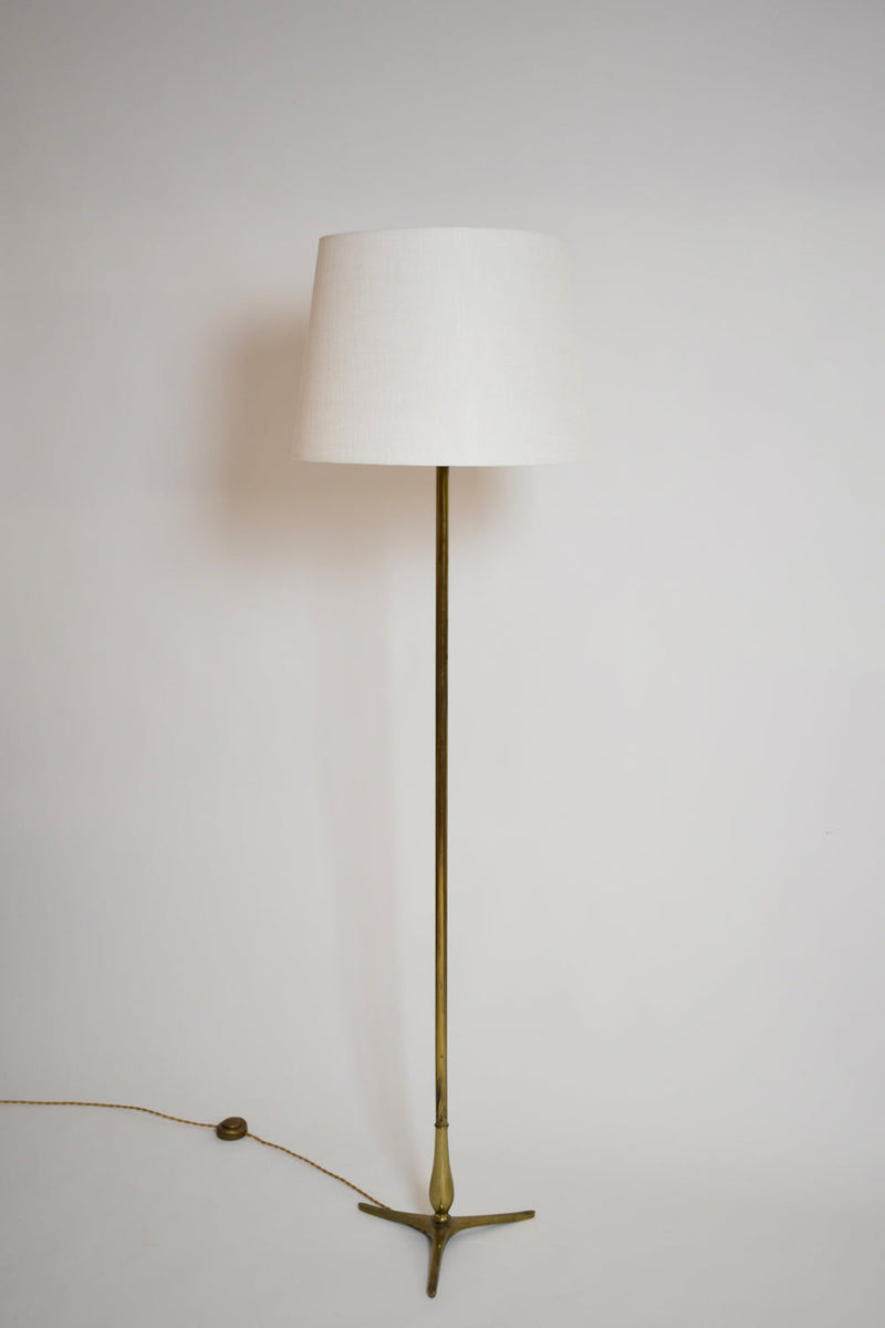 The Tripod Baluster Neoclassical Floor Lamp by Barracuda Interiors (1960s) features a slender brass stand, white tapered lampshade, and tripod base. It stands against a white background with an electric cord and foot switch extending from the base.