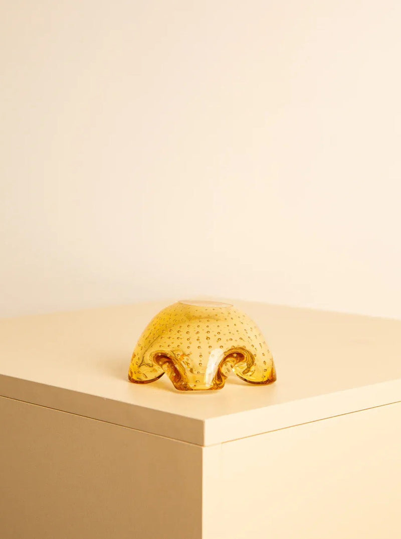 A small, translucent amber Treaptyque Vide-poches Murano 60's Yellow object with a textured surface and an organic, wave-like shape is placed on the corner of a light beige-colored cube. The matching beige background creates a minimalist aesthetic with a touch of retro design.