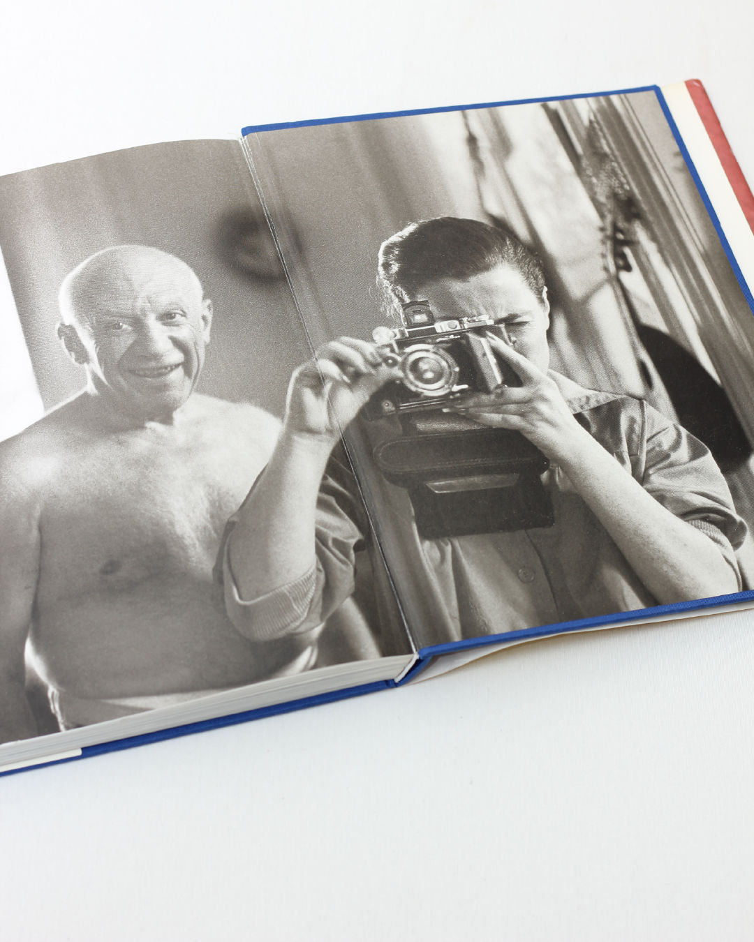 An open book, "Picasso and Jacqueline" from Boga Avante Shop, displays a black-and-white photo by David Douglas Duncan of a shirtless Picasso smiling beside someone holding a camera; the image spans two pages indoors.