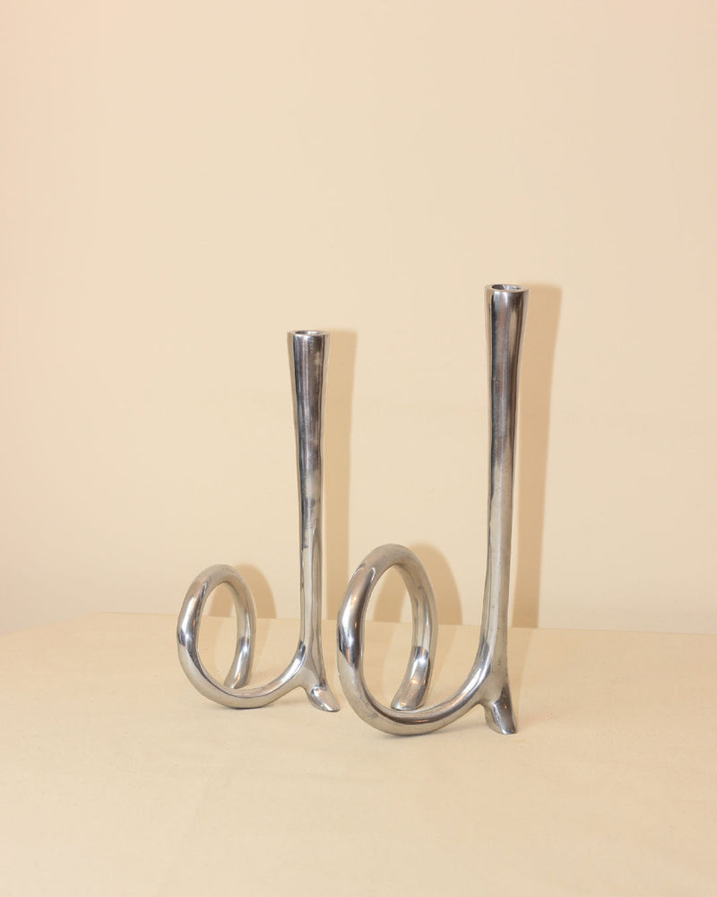 The Cast Aluminum Candleholders by Boga Avante Shop feature abstract, looped silver bases inspired by 1980s candelabras—ideal for modern minimalist decor, displayed on a beige surface with a light beige background.