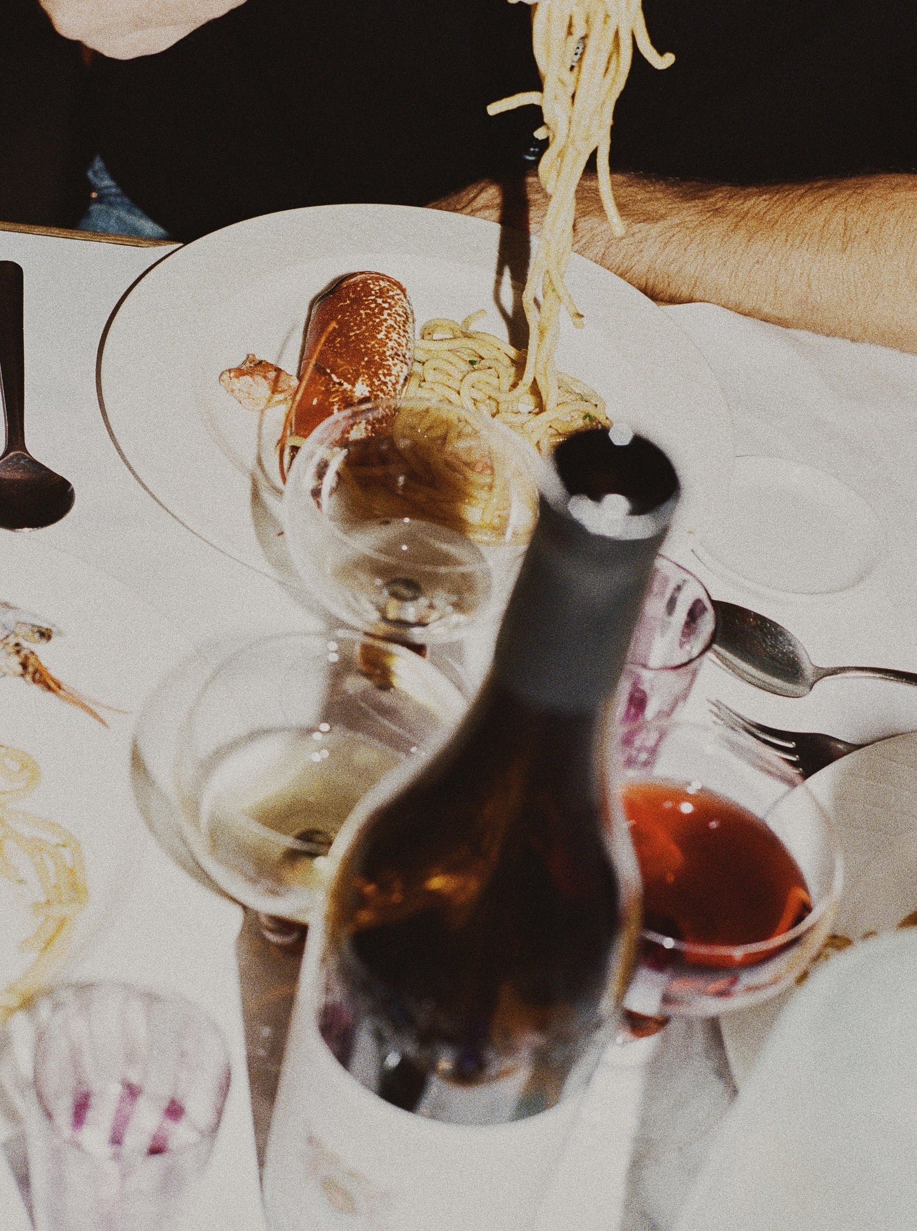 A dining table with a person's hand holding spaghetti above a plate, which also contains lobster. The table features Akua Objects' Noam Wine Glass - Set of Four, including a wine bottle, two empty glasses, and a glass of red wine. Other utensils are scattered around, and additional plates are partially visible, creating festive table settings.