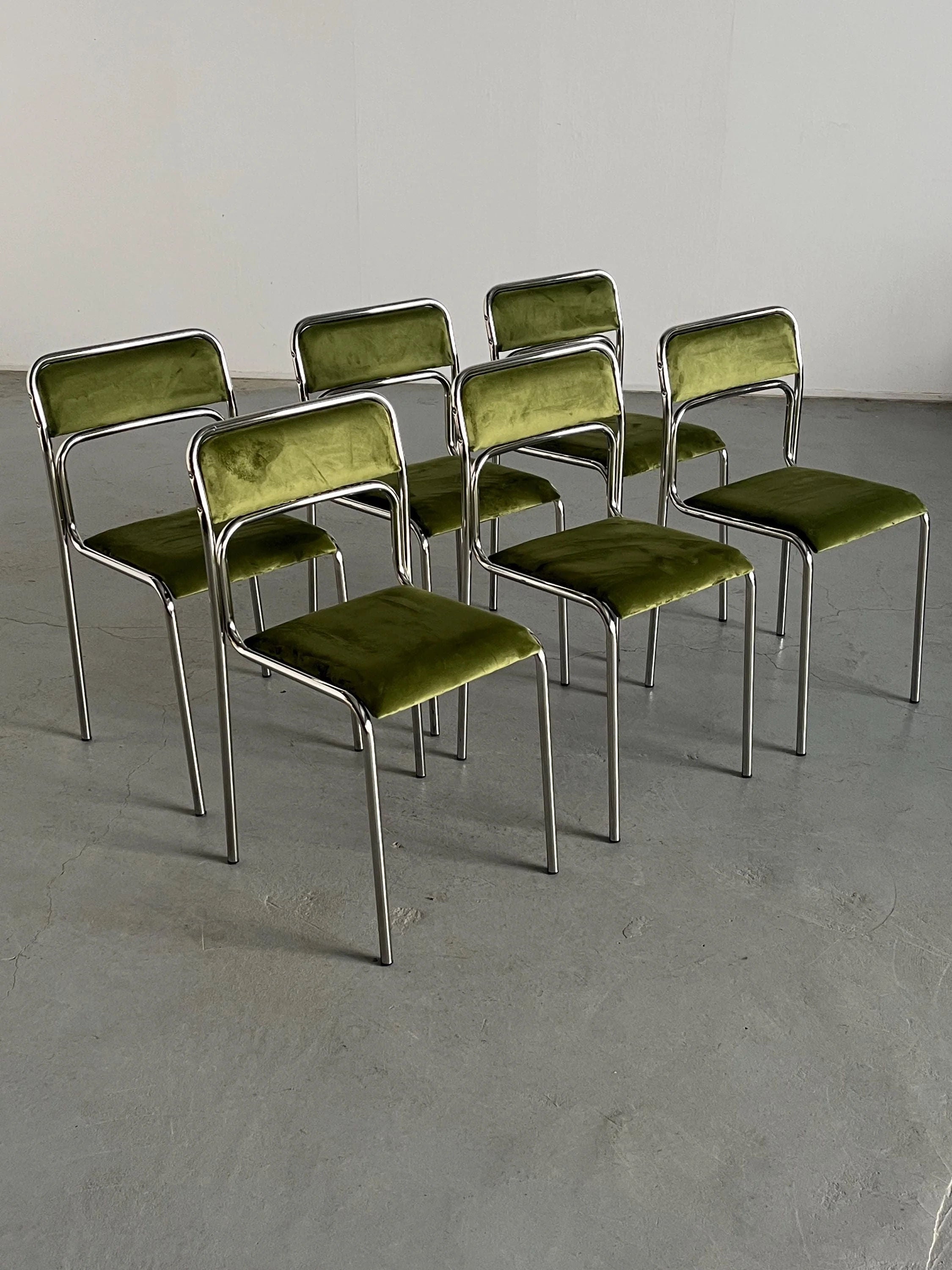 Six Cherry Cargo Bauhaus chrome and green velvet chairs are arranged in two slightly staggered rows on a gray concrete floor, against a plain light gray wall. Each chair showcases Italian modernist flair with its minimalist design featuring rectangular backrests and seats.